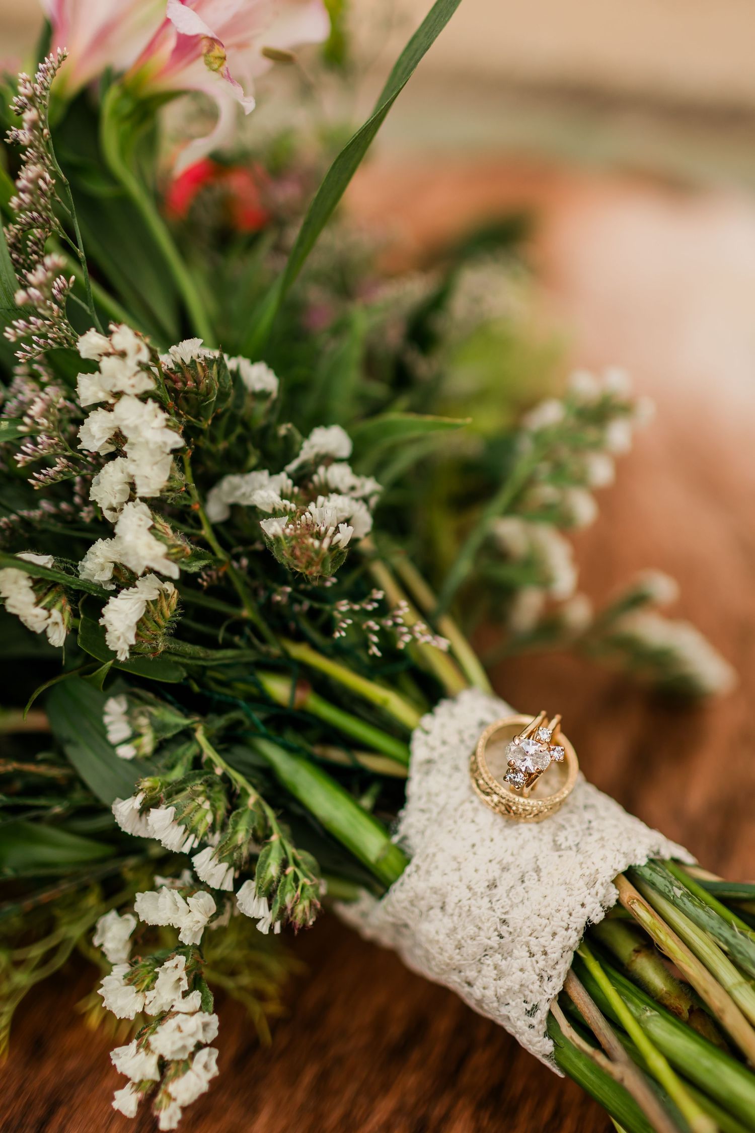 Gold wedding rings resting on a lace-wrapped bridal bouquet with white baby's breath and pink flowers.
