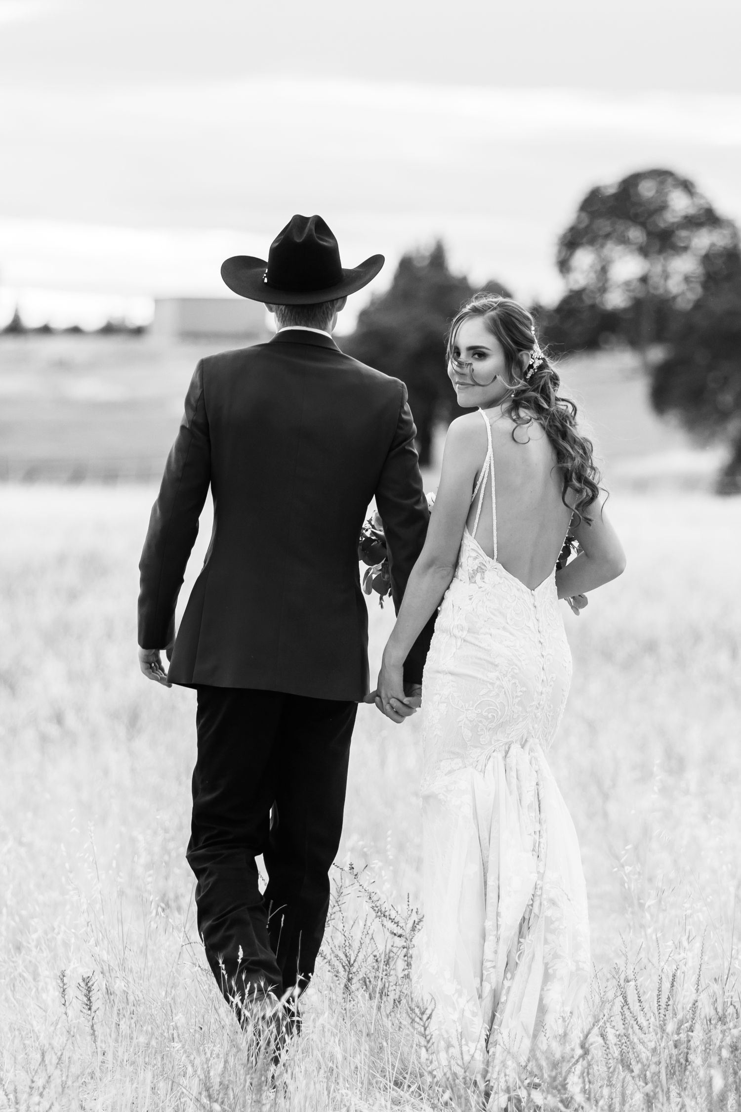 Black and white photograph of a couple walking through a meadow in wedding attire.