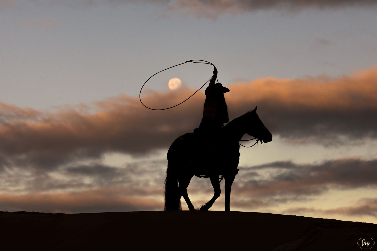 A silhouetted cowboy on horseback against a dramatic sunset sky with orange clouds and glowing sun.