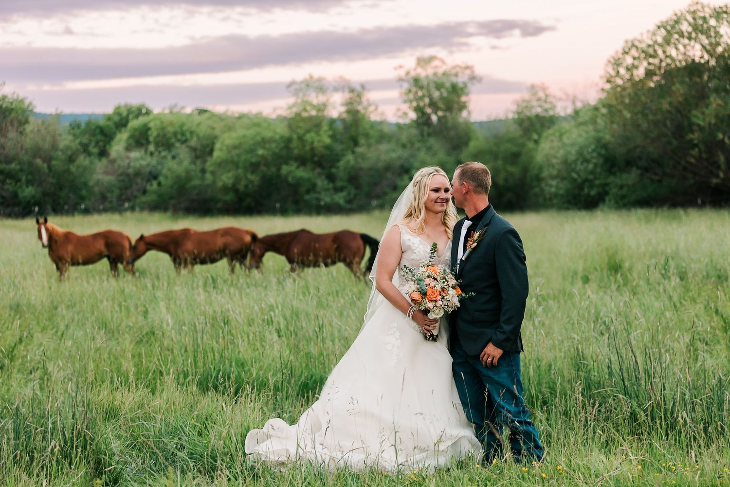 A romantic wedding photo session in a green meadow with brown horses grazing in the background.