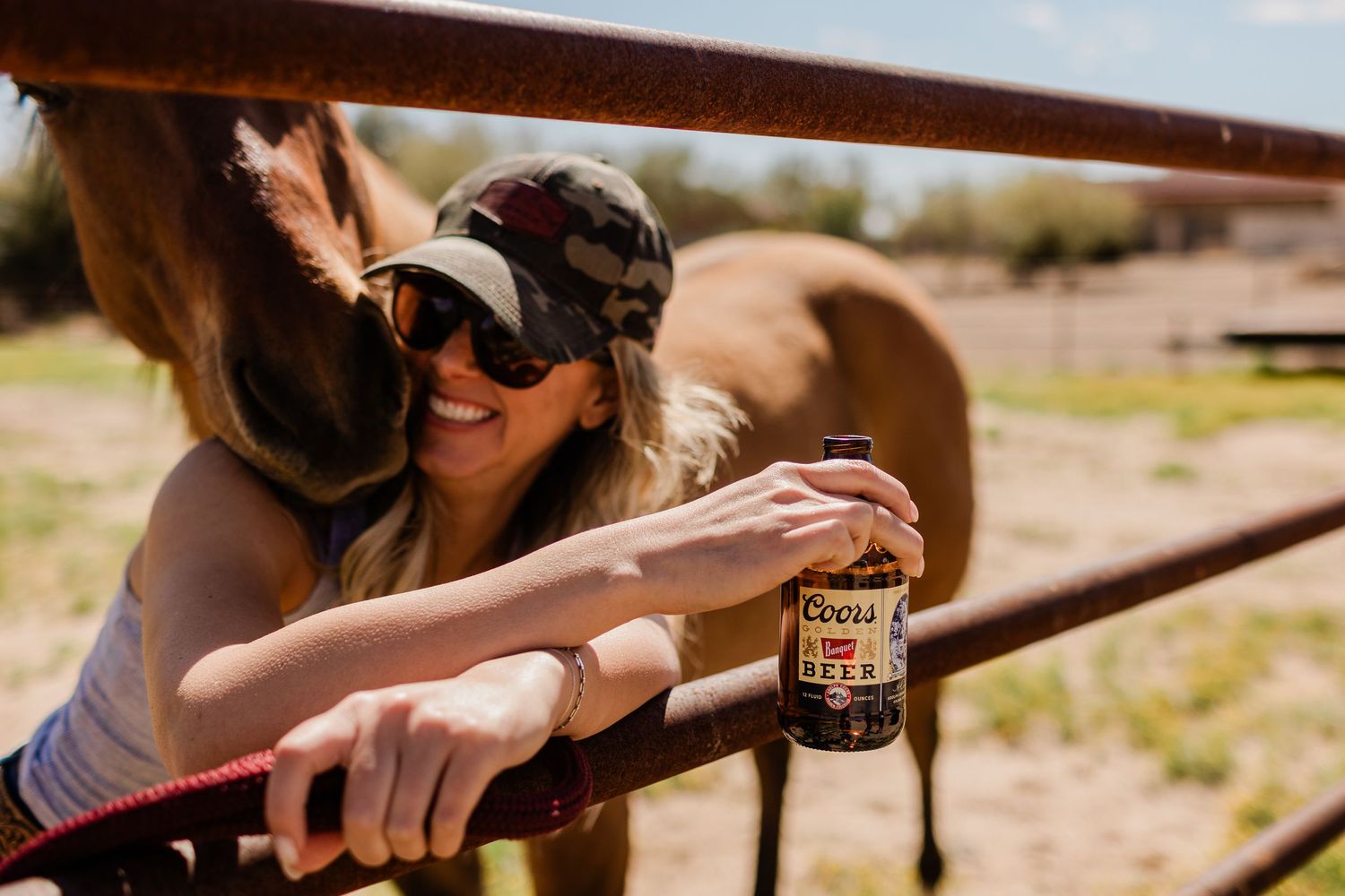 Hand holding beverage bottle near brown horse at wooden fence rail.
