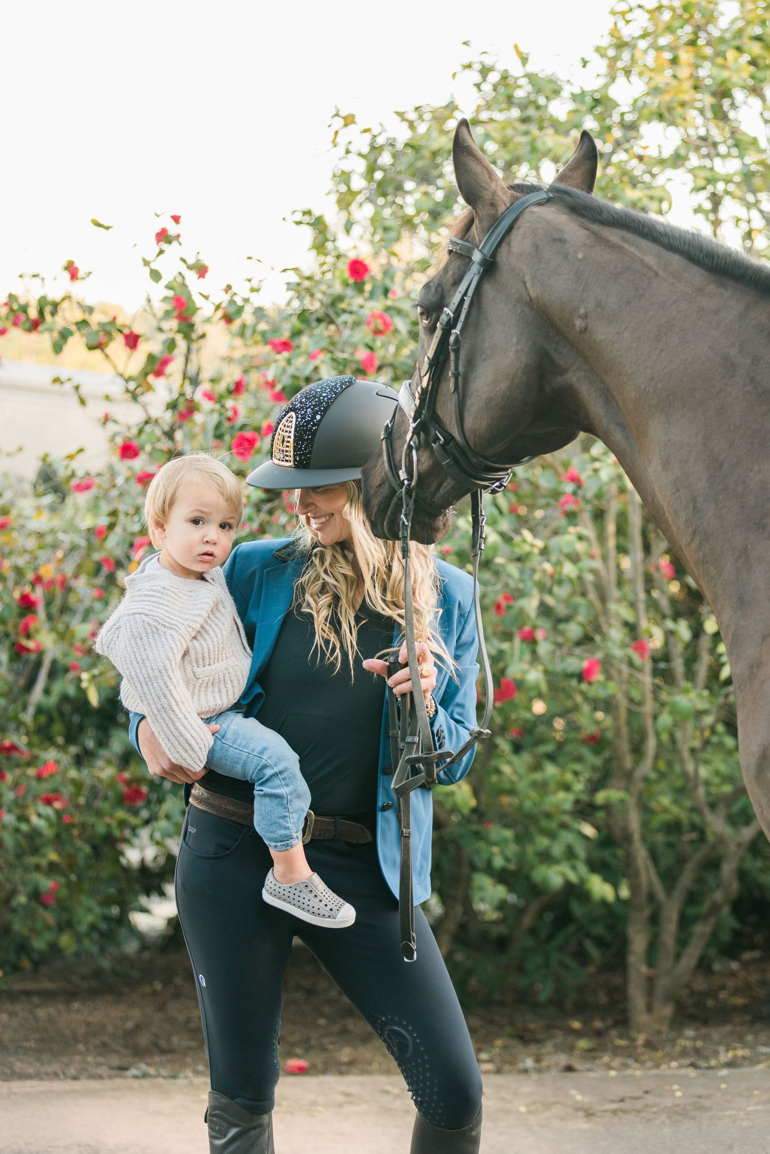Horse with black coat stands near blooming rose garden on sunny day.