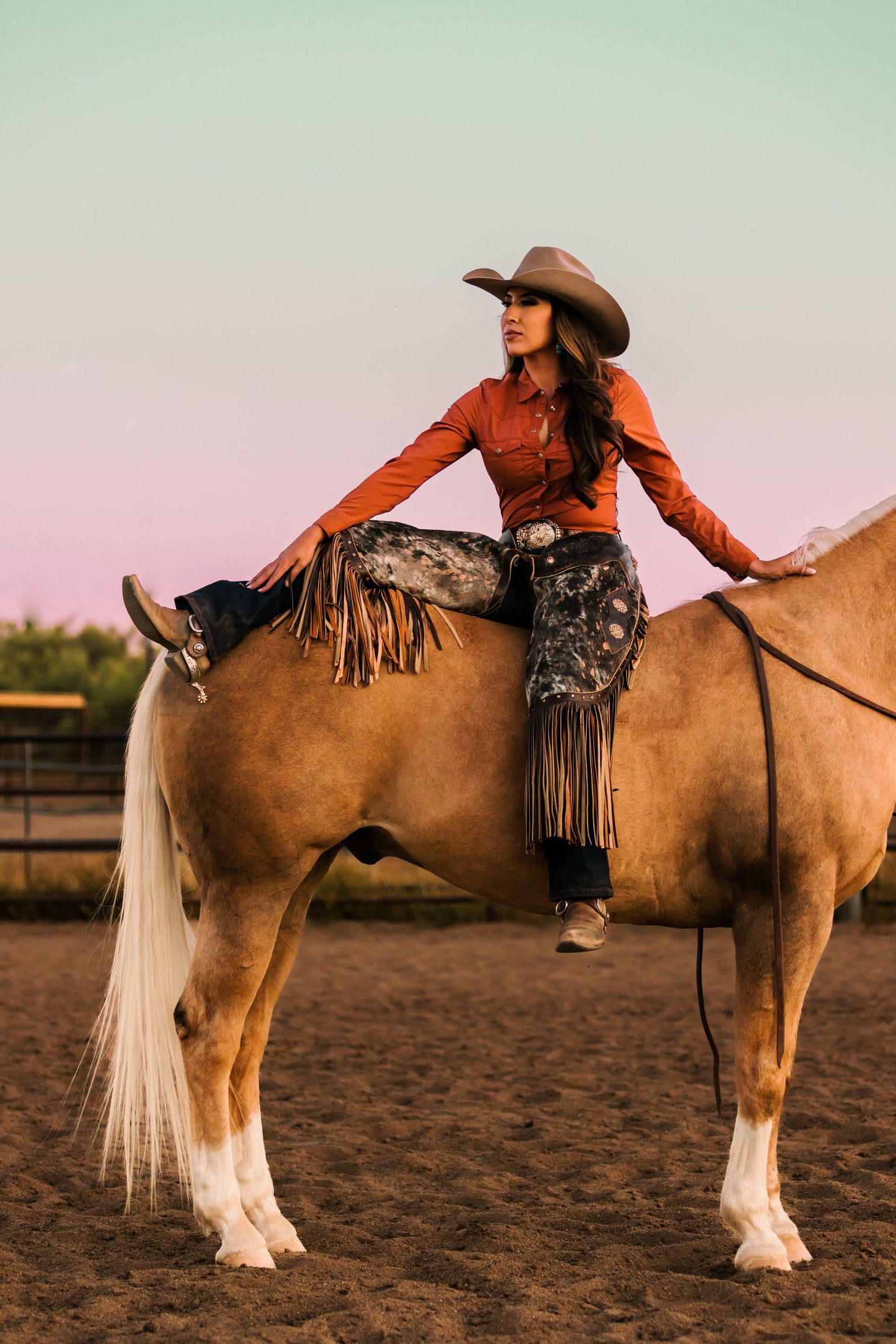 Rider in orange shirt and cowboy hat performs stretching exercises while mounted on a palomino horse in dusty arena.