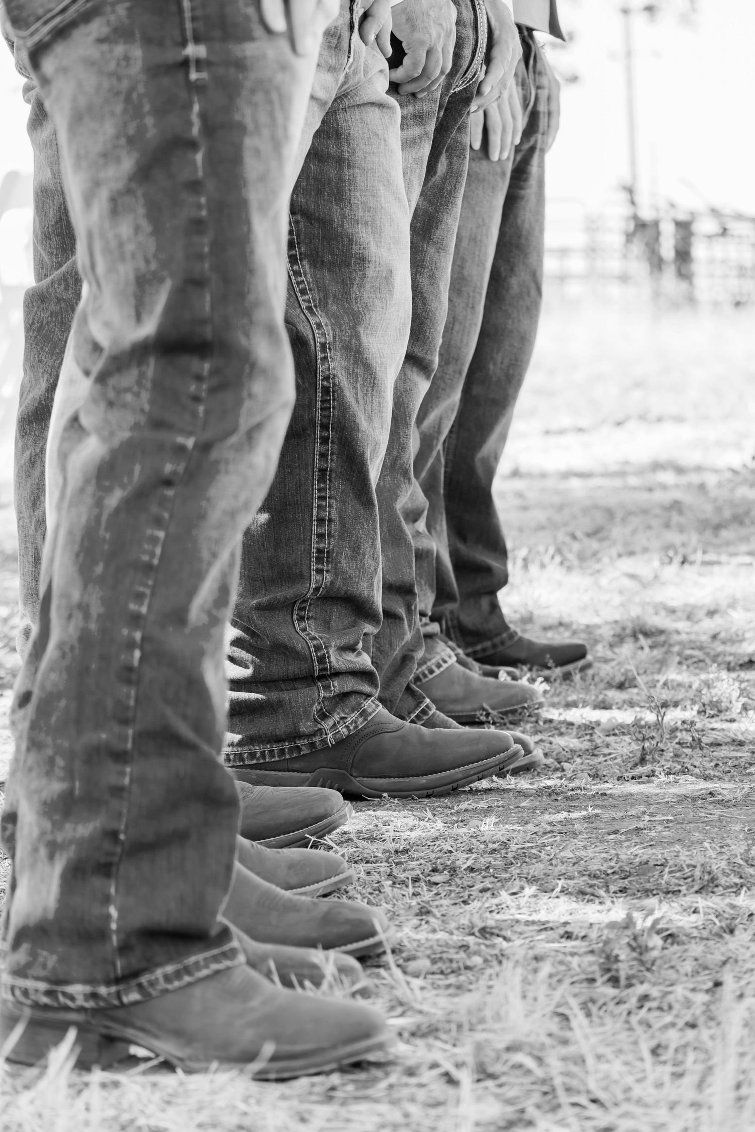 A black and white view of people's legs wearing jeans and work boots standing in a line on dirt ground.