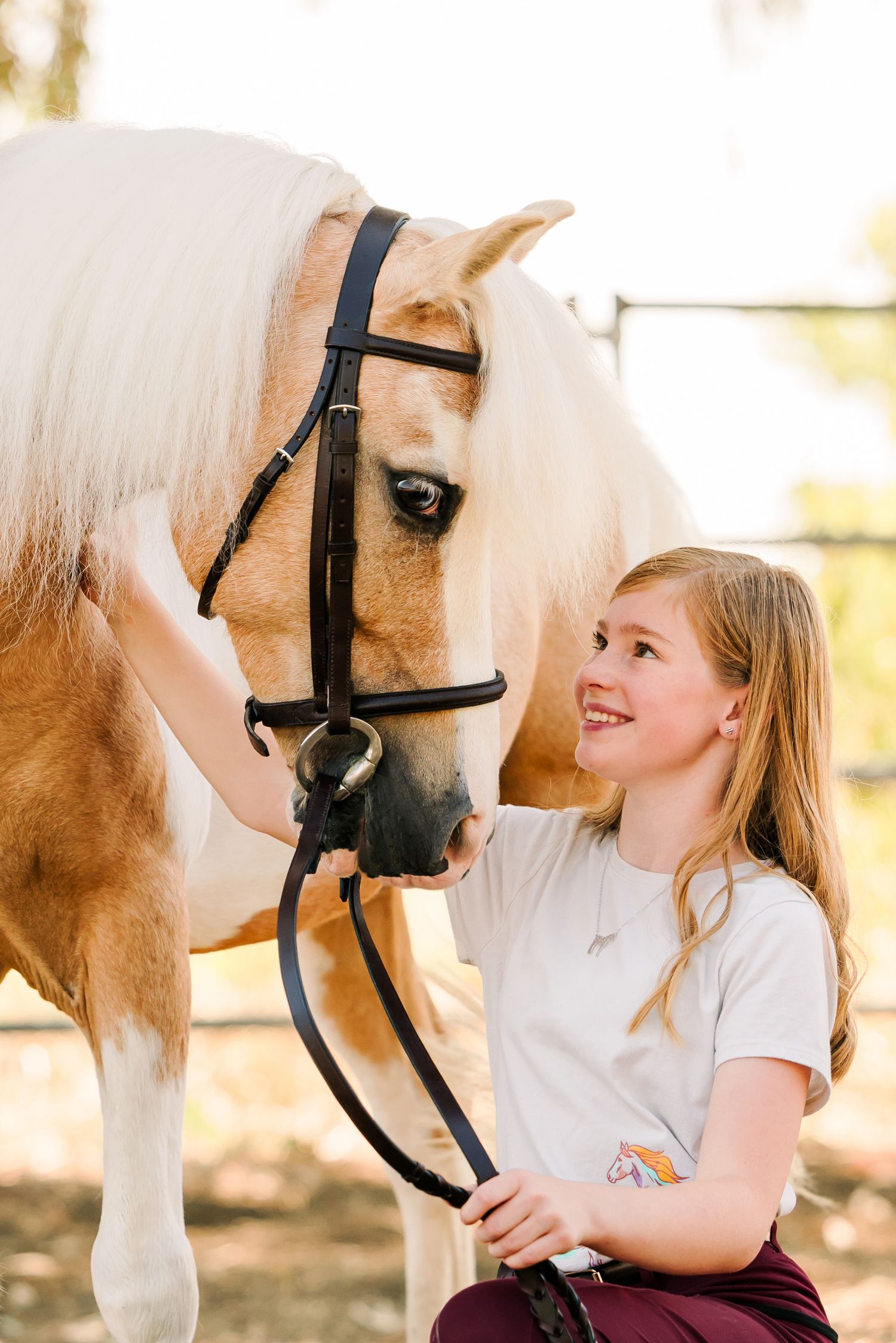 Young girl in white shirt lovingly interacts with a cream-colored horse wearing a black bridle.