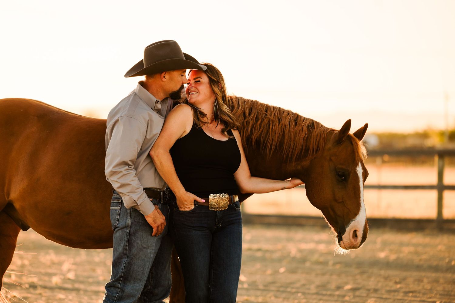 A romantic couple poses with their horse during a golden sunset photoshoot in a rustic outdoor setting.
