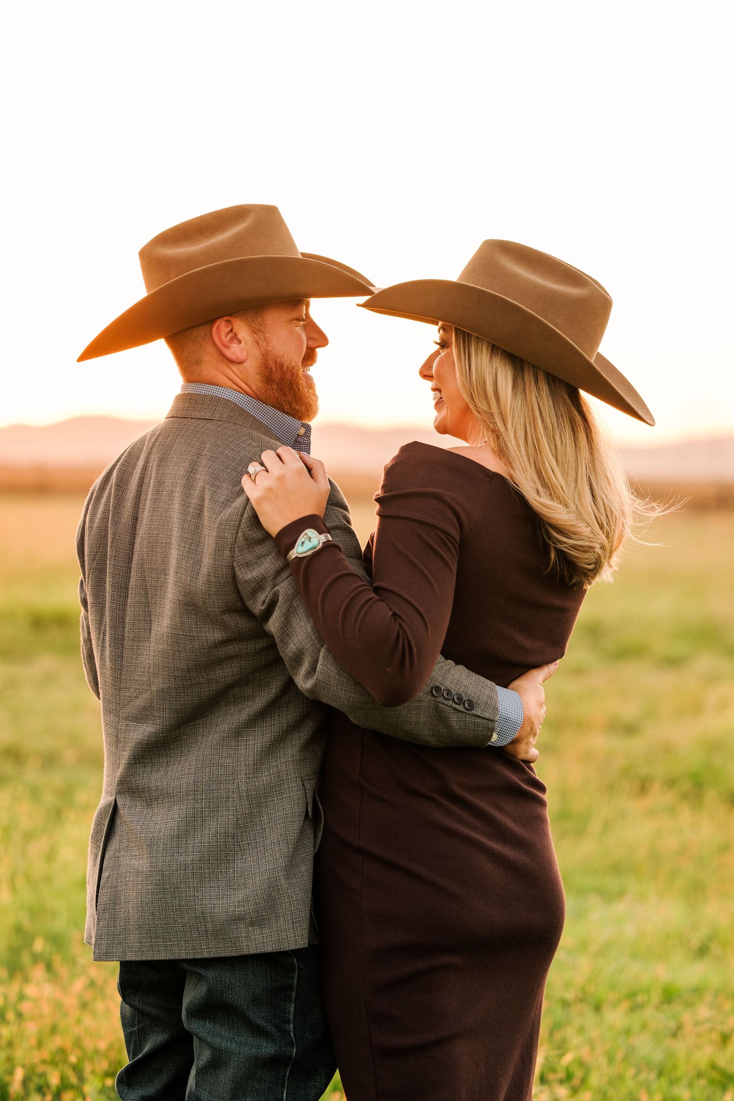 A couple in cowboy hats embrace in a field at sunset wearing western attire and formal clothing.