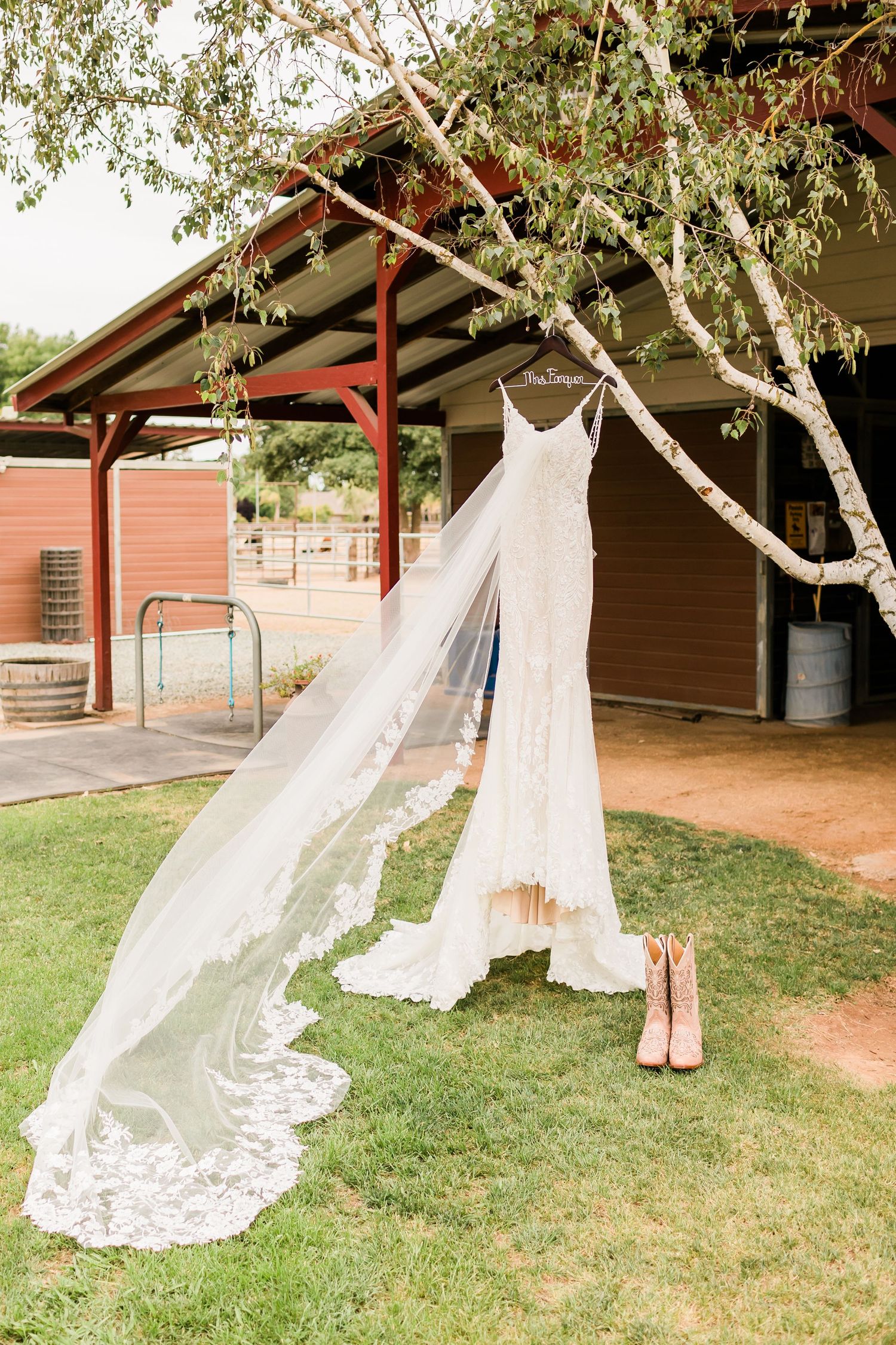 Wedding gowns and pink heels hang delicately from tree branches in an outdoor rustic setting.