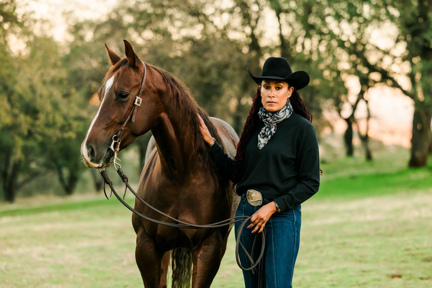 A brown horse and rider stand together in a grassy field during a photo session at sunset.