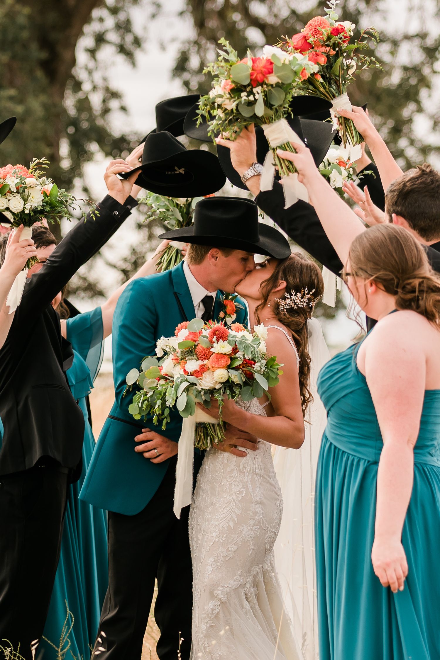 Wedding party holding black cowboy hats up while couple shares kiss surrounded by teal bridesmaid dresses.