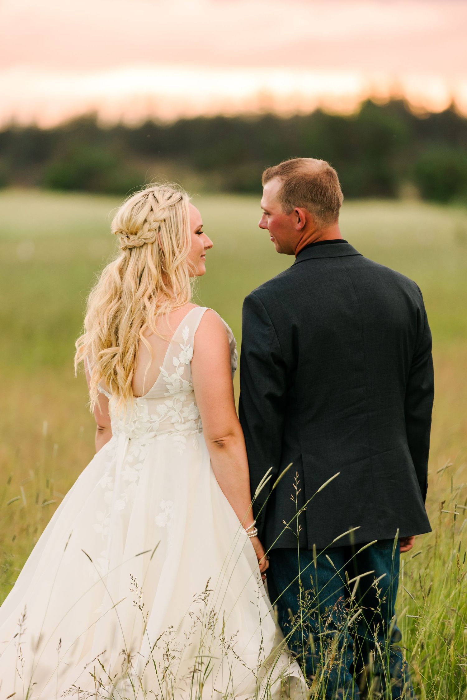 Bride in white lace gown and groom in black suit standing together in sunlit field of tall grass at sunset.