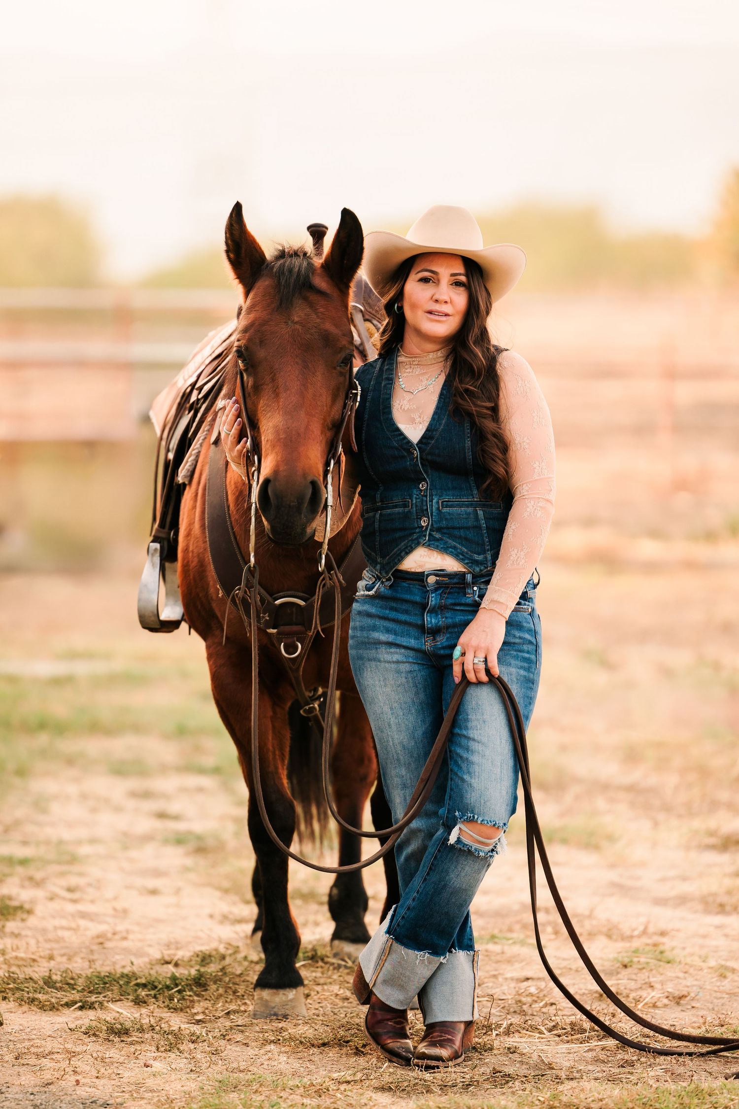 A brown horse stands beside someone wearing jeans and boots during a golden sunset photoshoot.