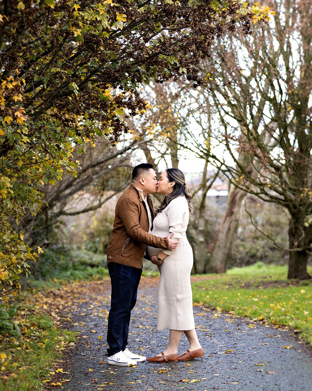 A couple shares a tender moment on a leaf-covered path surrounded by autumn trees.