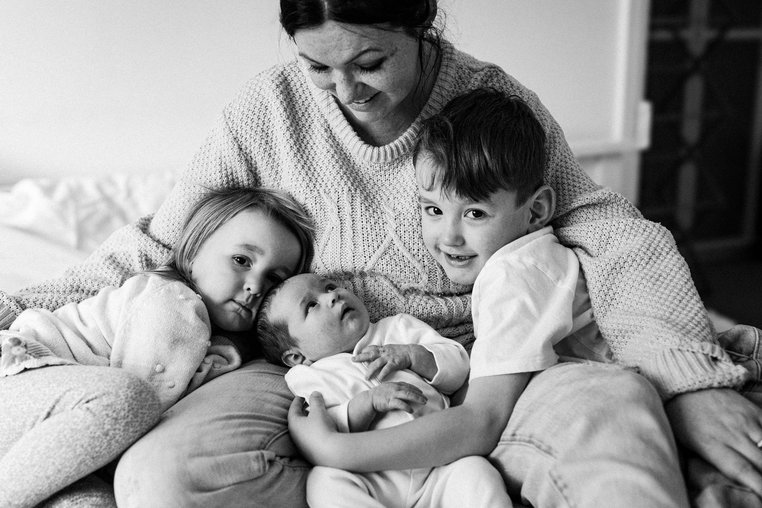 A loving black and white photograph of a parent and three small children cuddled together and smiling on a bed.