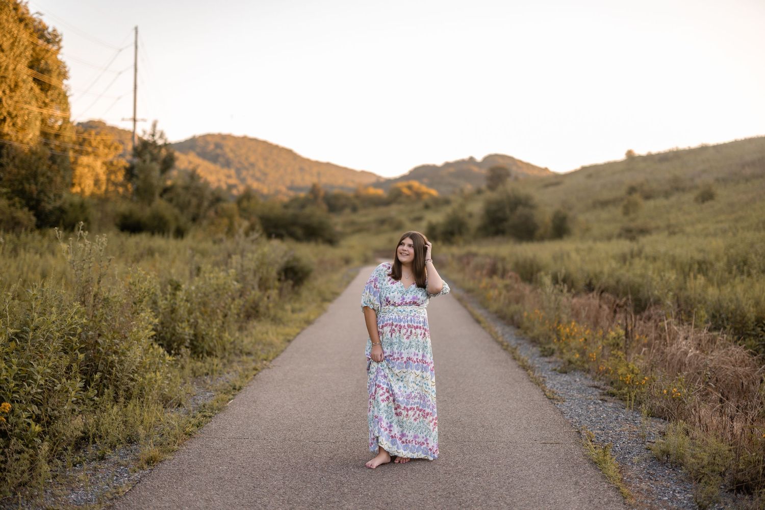 Someone in a floral maxi dress stands on a dirt path through golden hills at sunset.