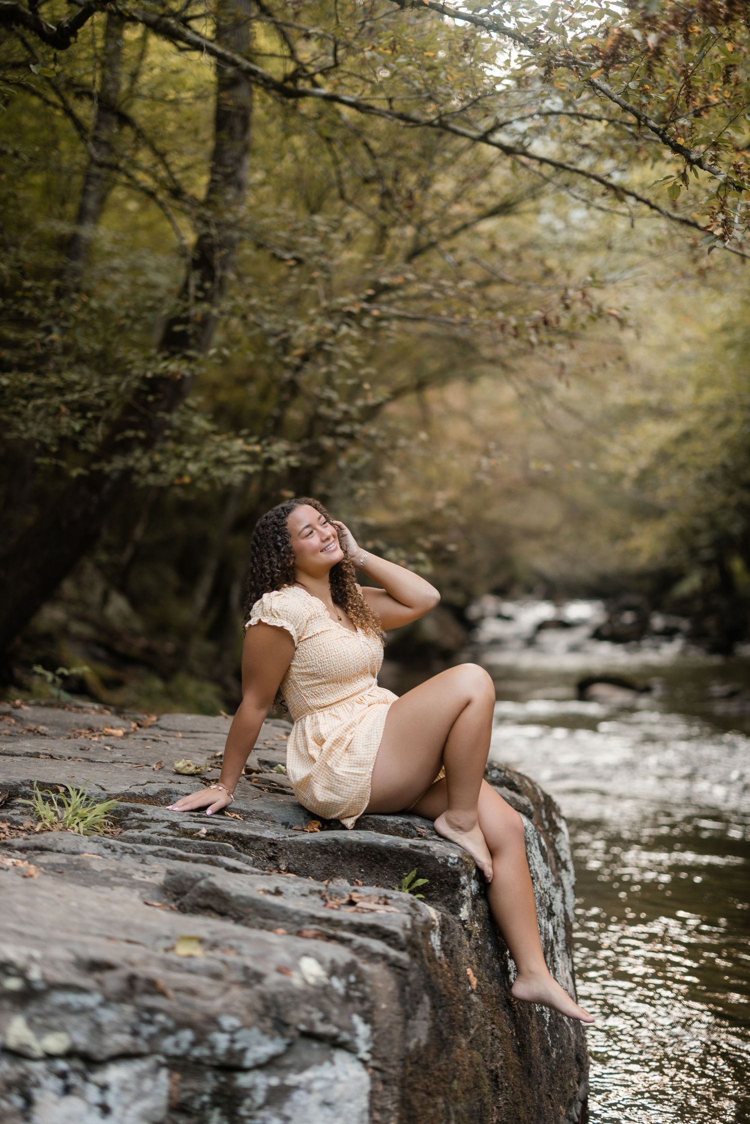 A person in a white dress poses gracefully on rocks beside a flowing stream in an autumn forest setting.