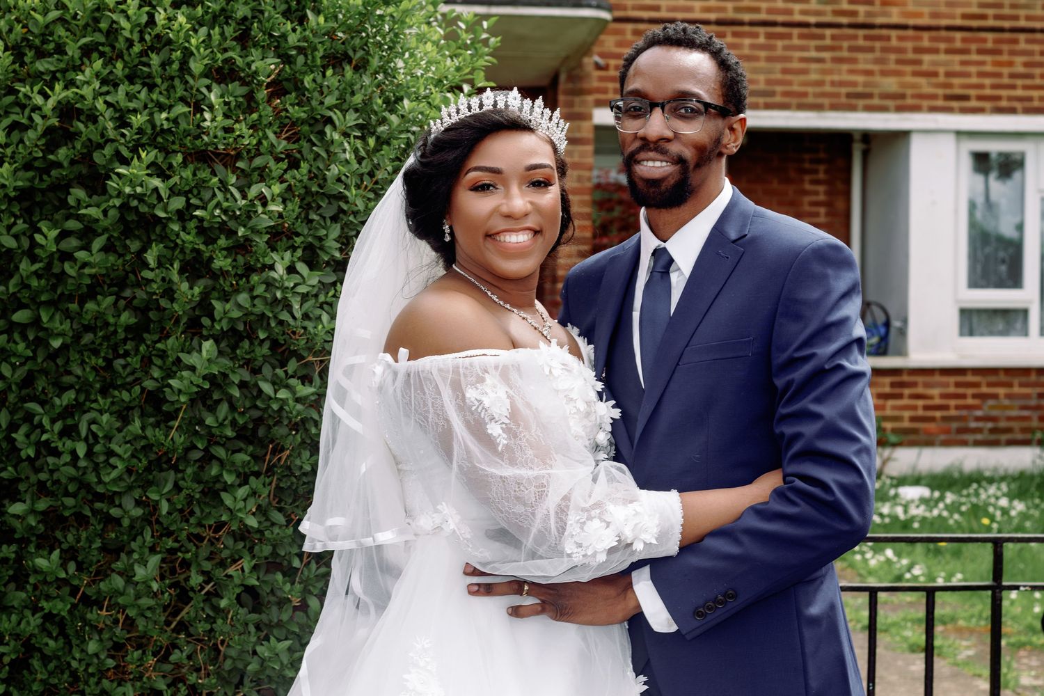 A newlywed couple embraces outdoors in front of a brick building, wearing a white wedding gown and navy suit.