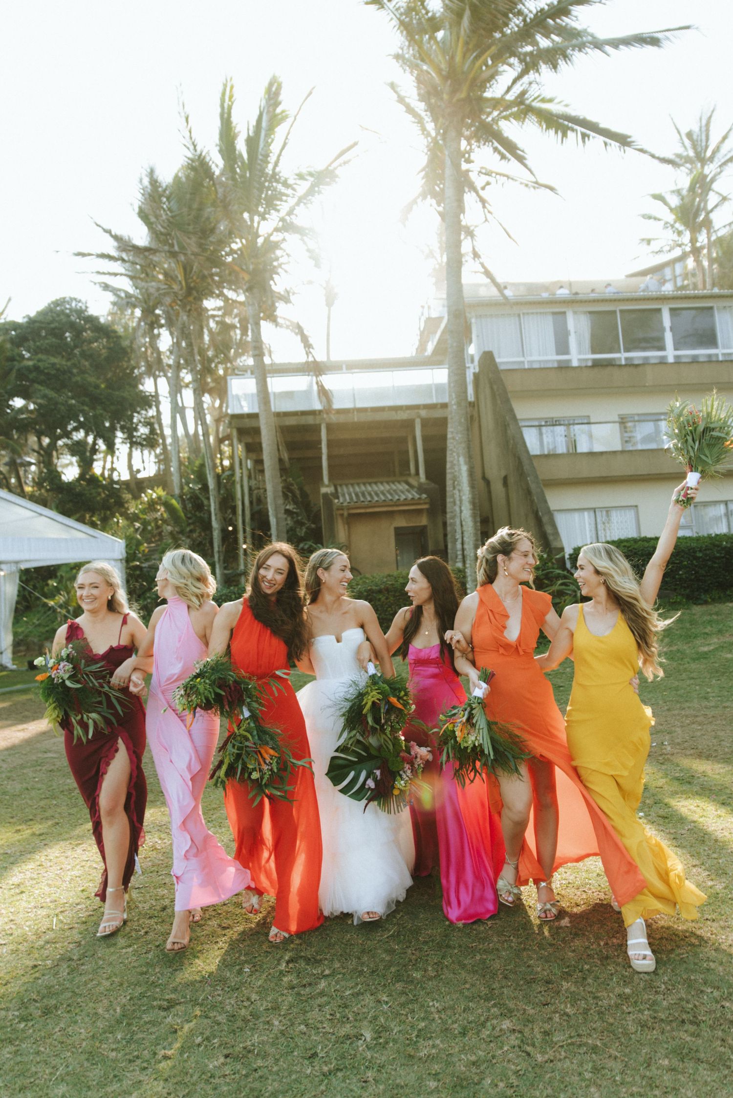 A bridal party in vibrant rainbow-colored dresses poses together on a grassy lawn with palm trees at sunset.