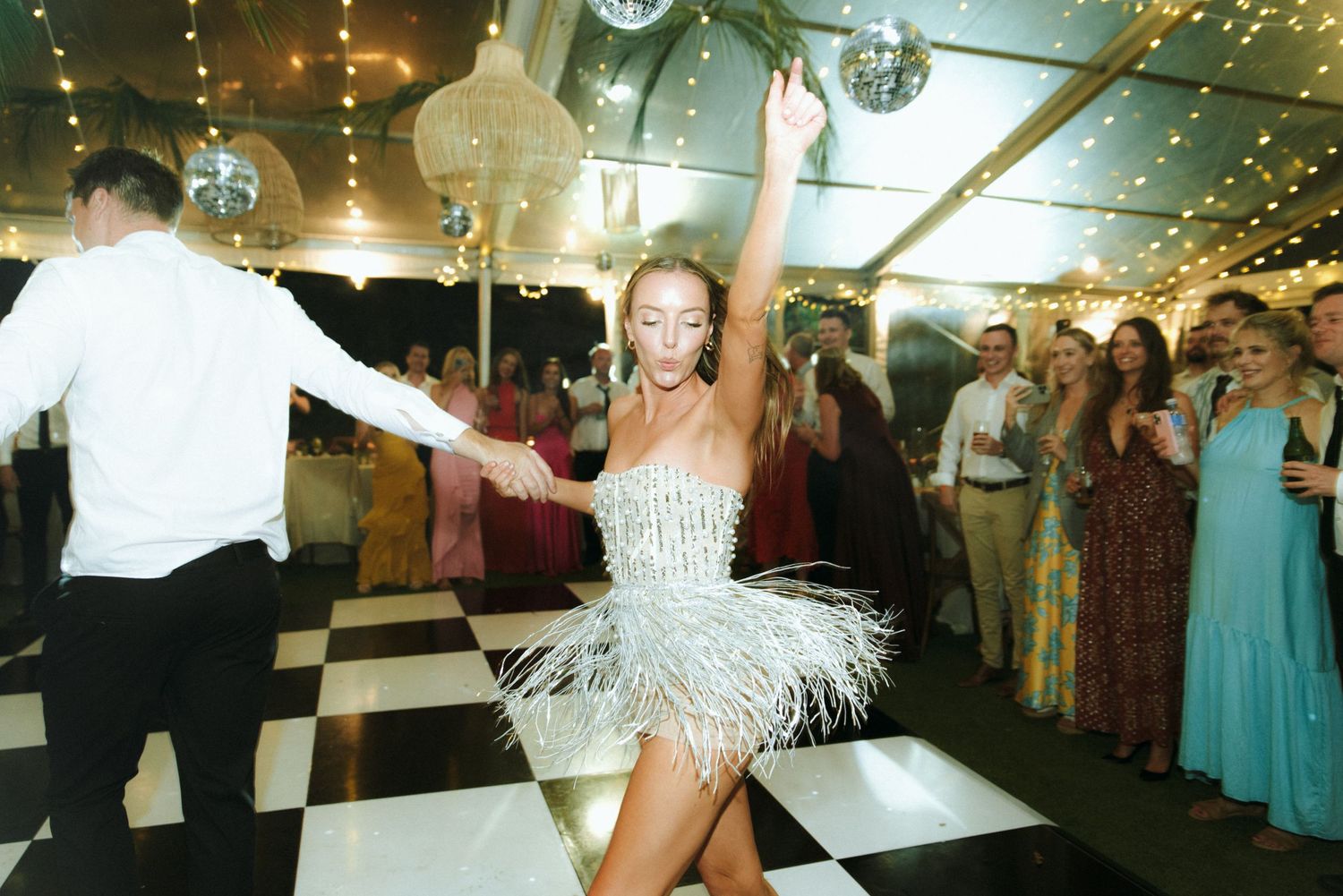 Wedding guests dance and celebrate under twinkling lights and disco balls at an indoor reception venue.