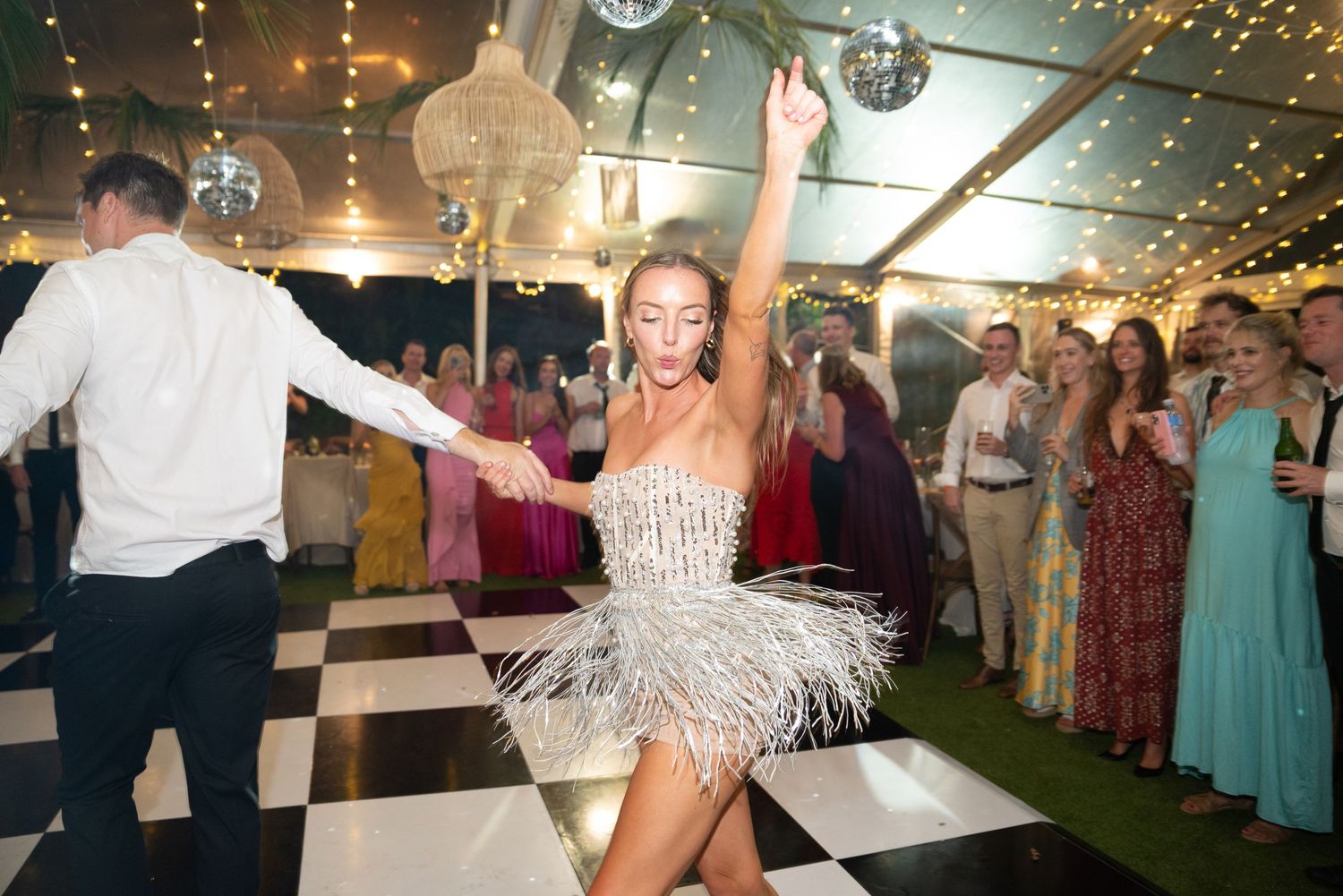 People dancing and celebrating at a wedding reception under disco balls and string lights in a tent venue.