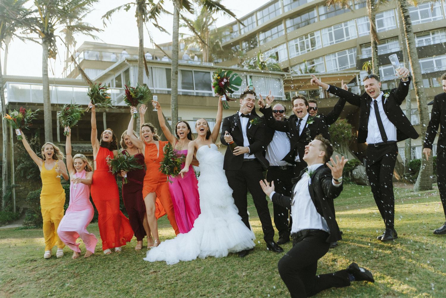 Wedding party in formal attire jumps joyfully on grass with palm trees and buildings in the background.