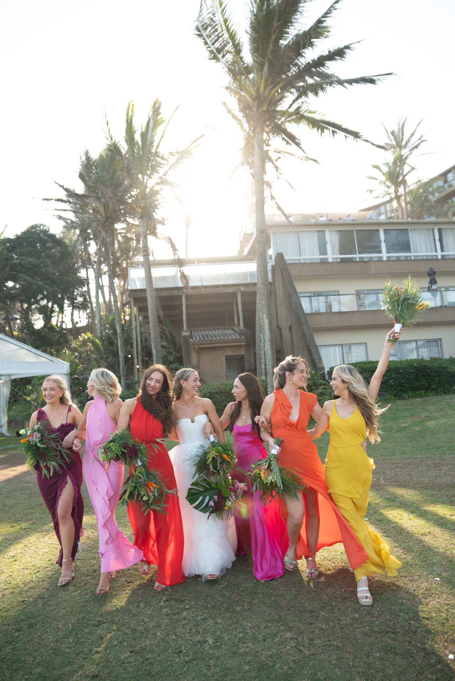 A group in colorful formal dresses poses together on a grassy lawn with palm trees at sunset.