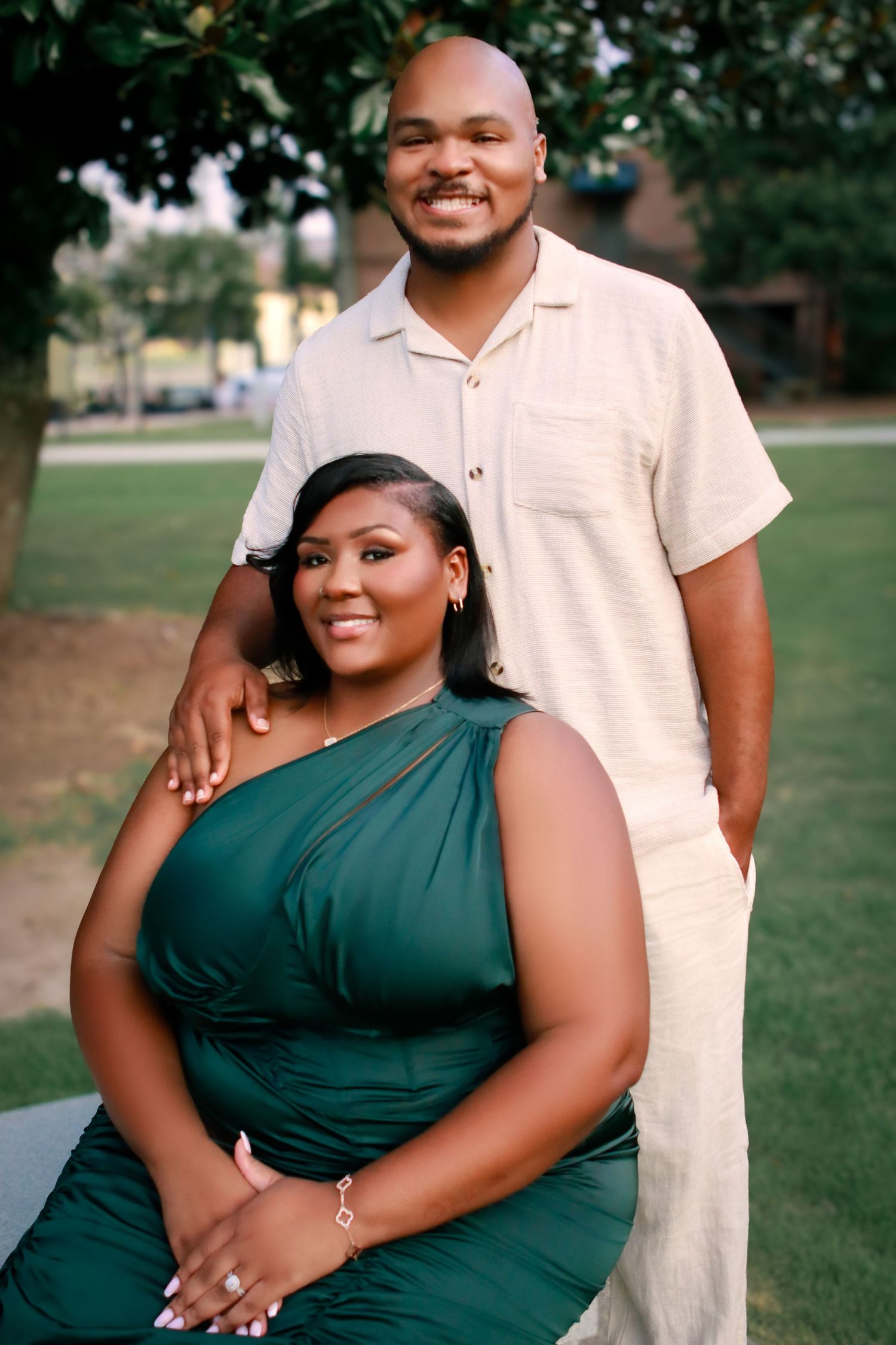A couple poses together outdoors on green grass, with one person standing and one sitting.