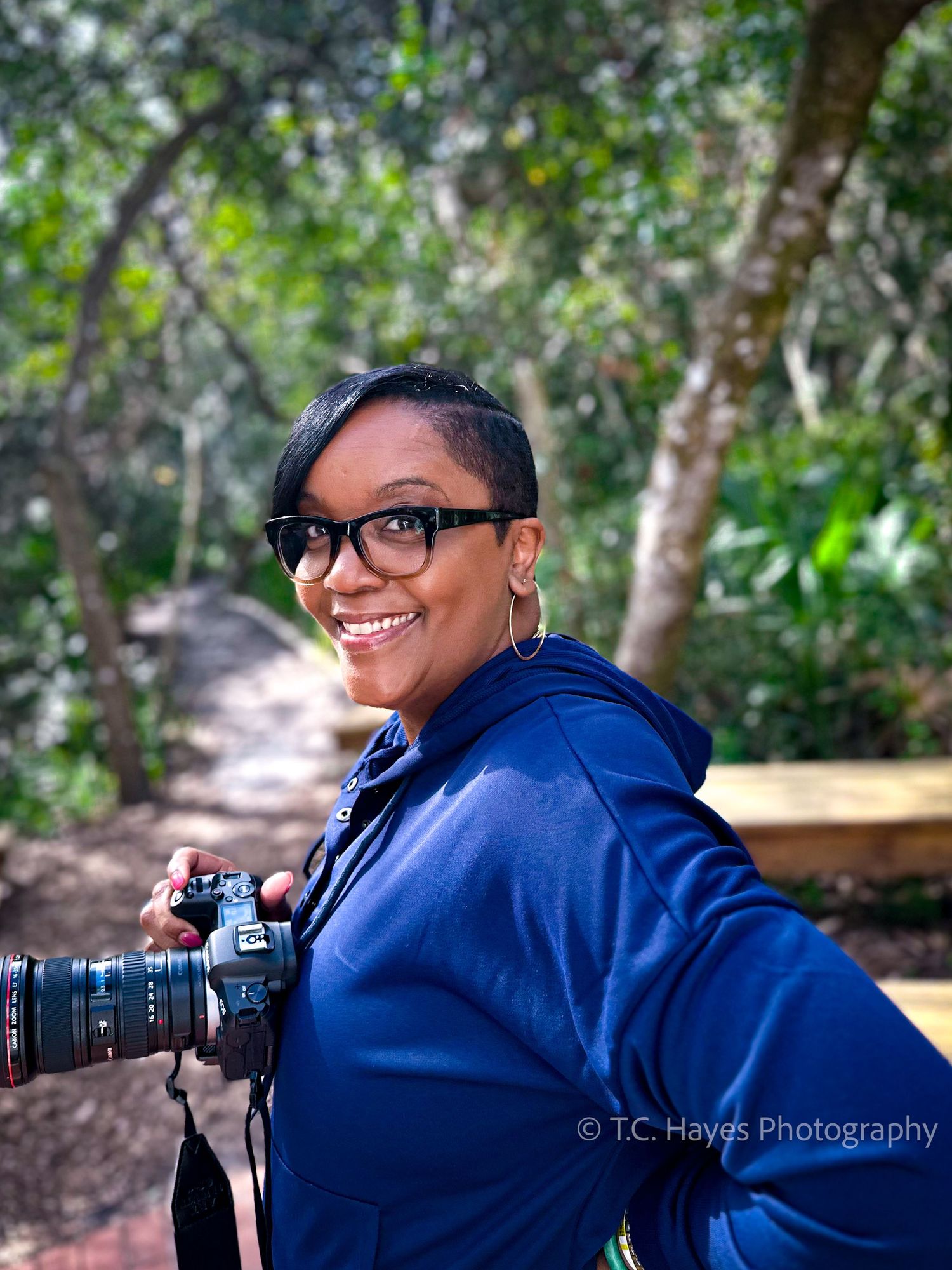 A photographer wearing a blue hoodie and glasses holds a professional camera outdoors.