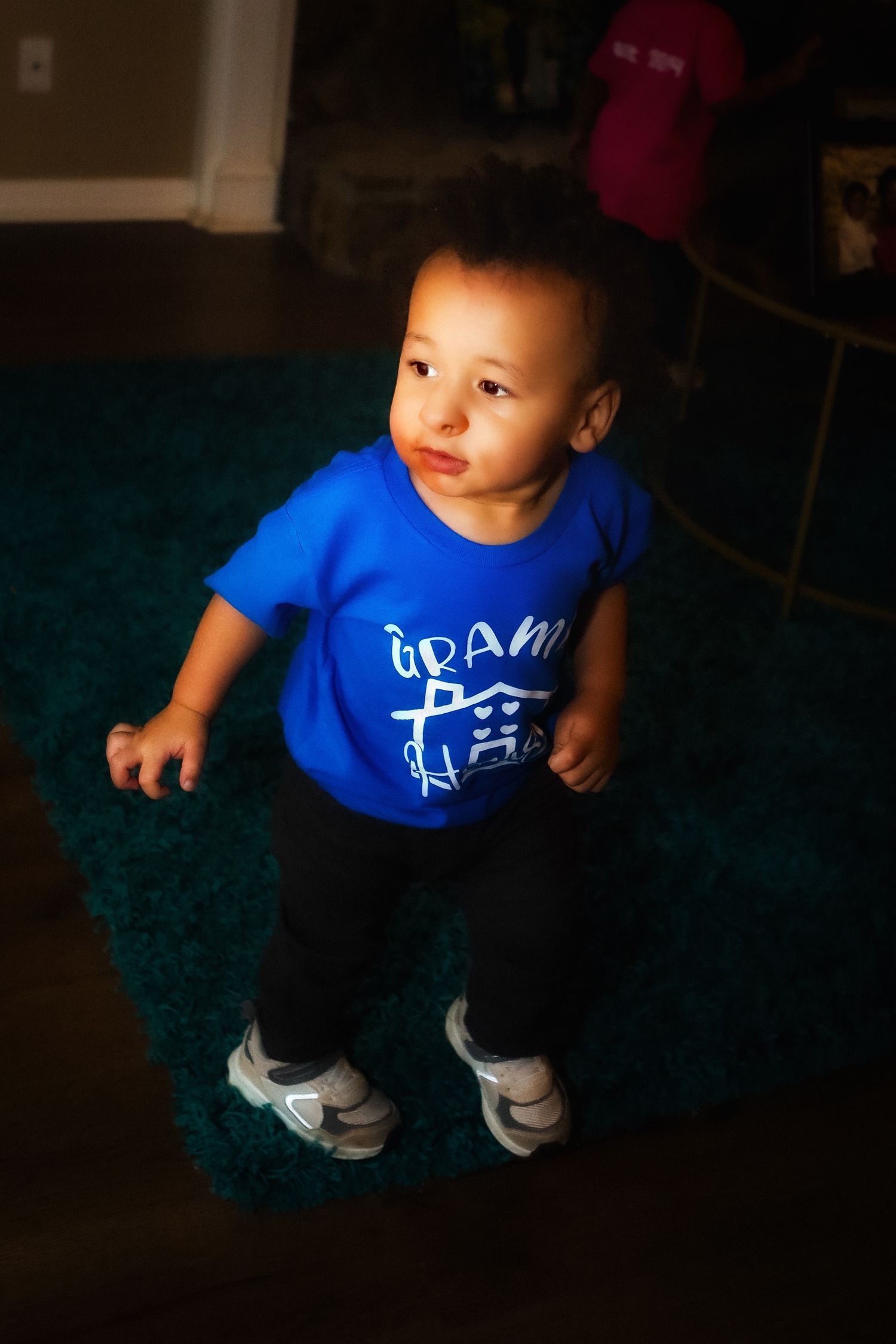 Young child in a bright blue t-shirt stands on a dark carpet in low lighting.