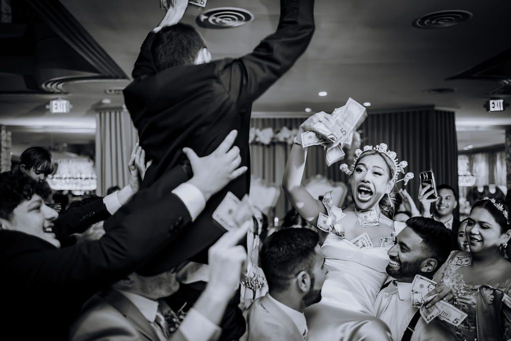A joyous wedding celebration captured in black and white as guests lift and cheer during a lively reception.