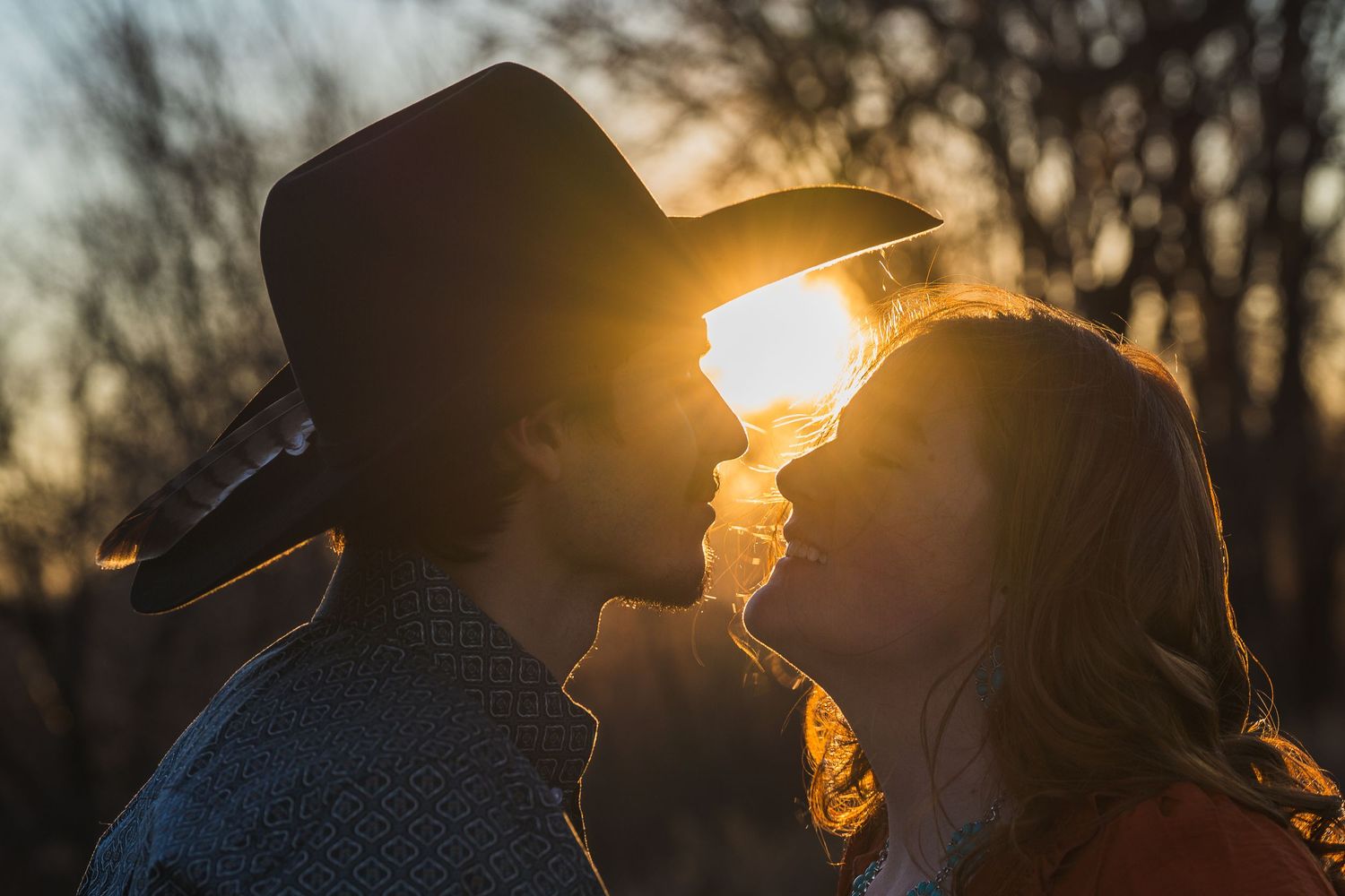 A silhouetted couple shares an intimate moment at sunset with sun rays peeking through between them.
