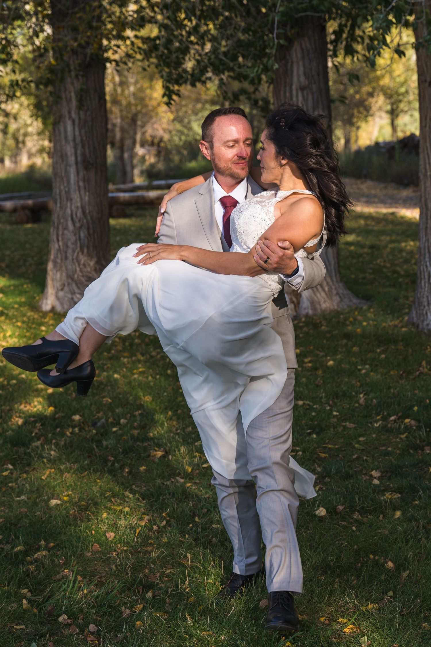 A couple in white outfits dances playfully on grass with trees in the background during a sunny autumn day.