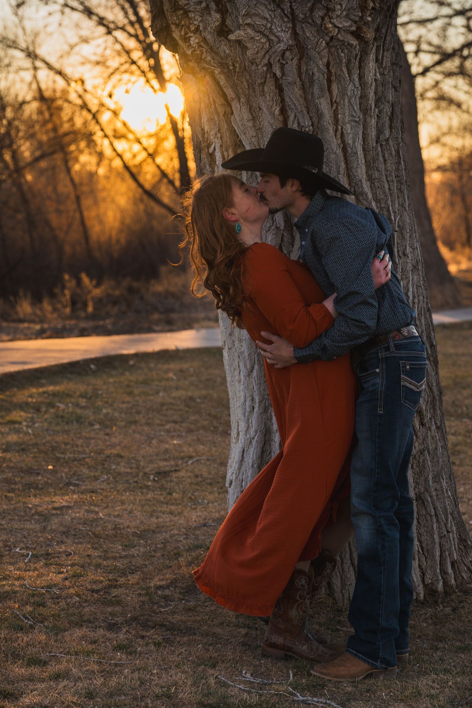 A couple in denim and rust-colored clothing share an intimate embrace against a tree during golden hour sunset.