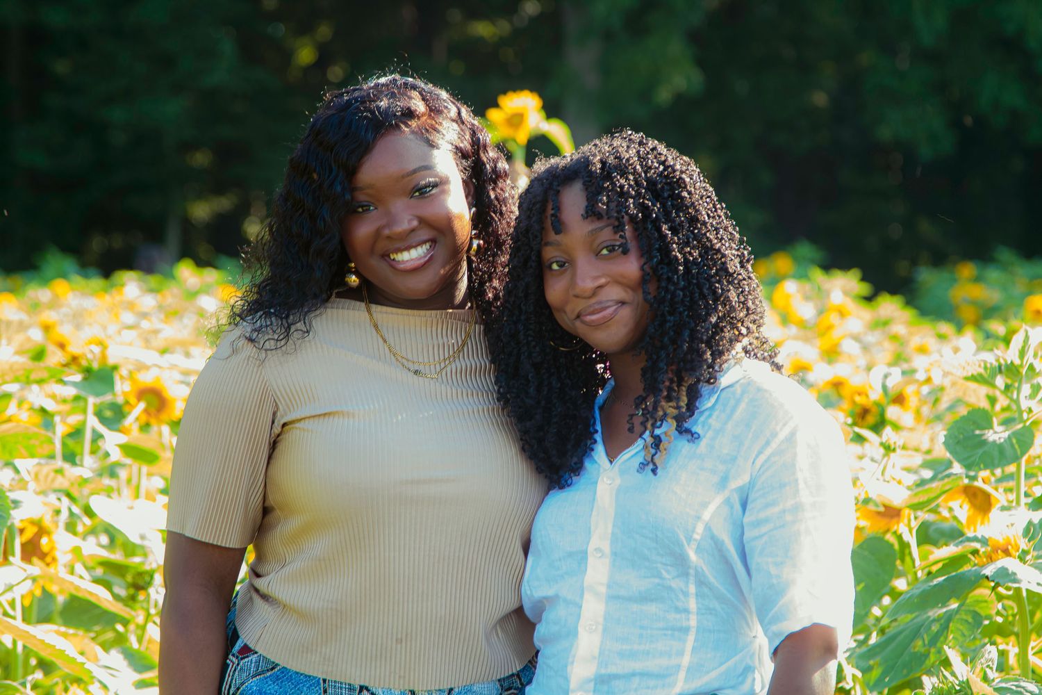 Two women smiling together in a sunflower field, wearing casual outfits on a sunny day.