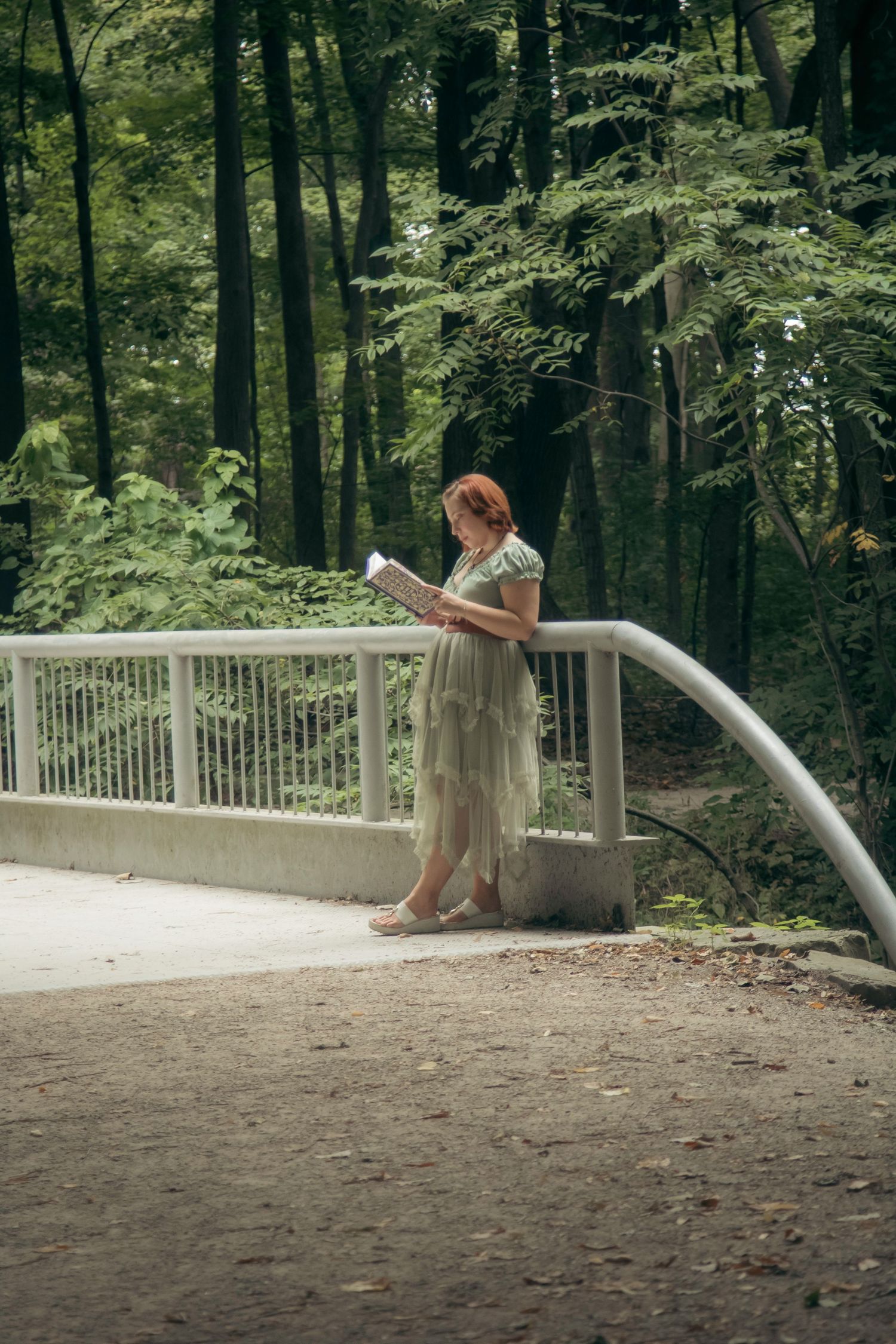 Woman in a flowing sage green dress reads a book while leaning on a white bridge railing, surrounded by lush green forest.