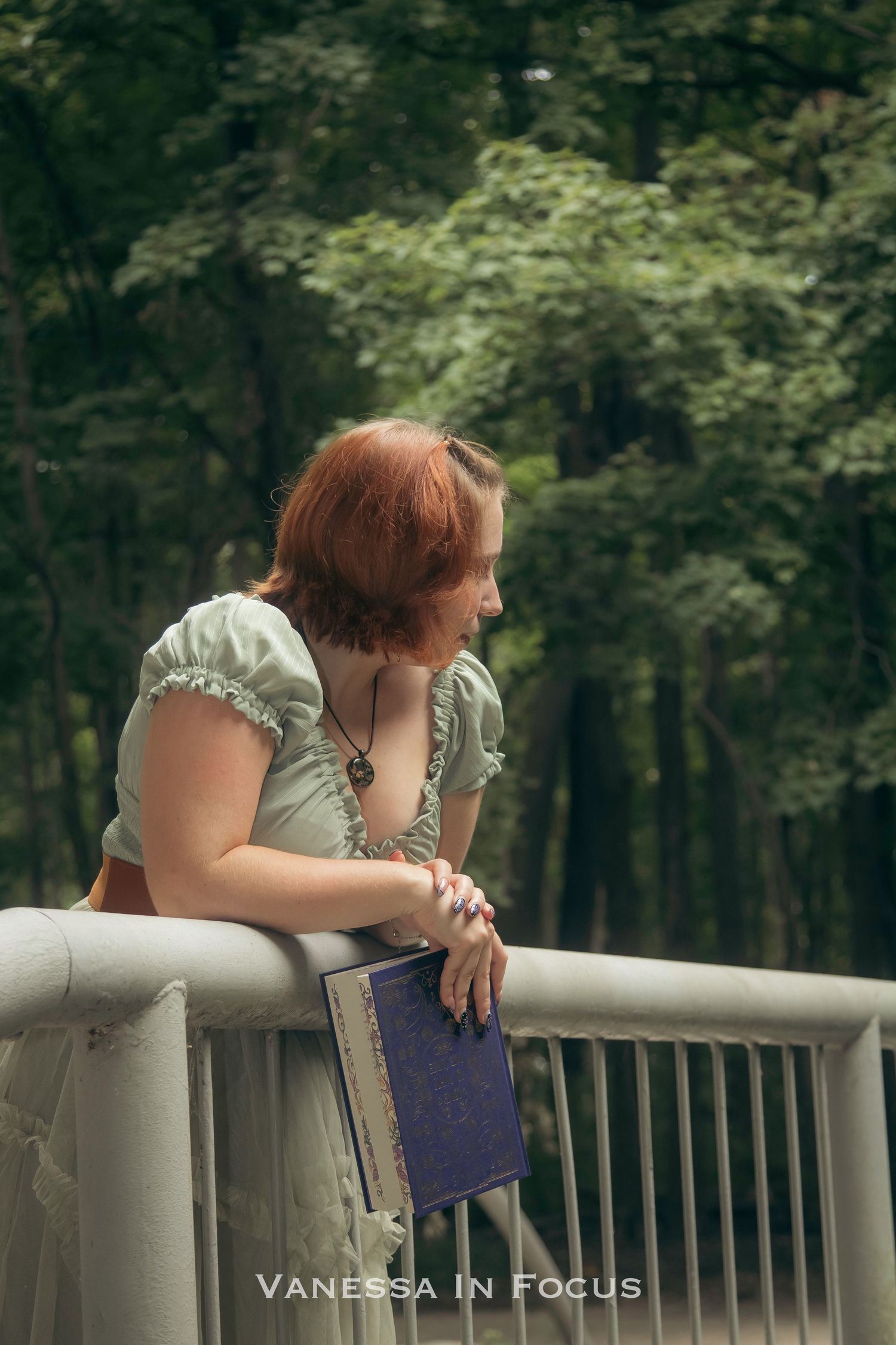 A red-haired woman in a mint dress leans on a bridge railing, holding a book, gazing into a lush green forest.