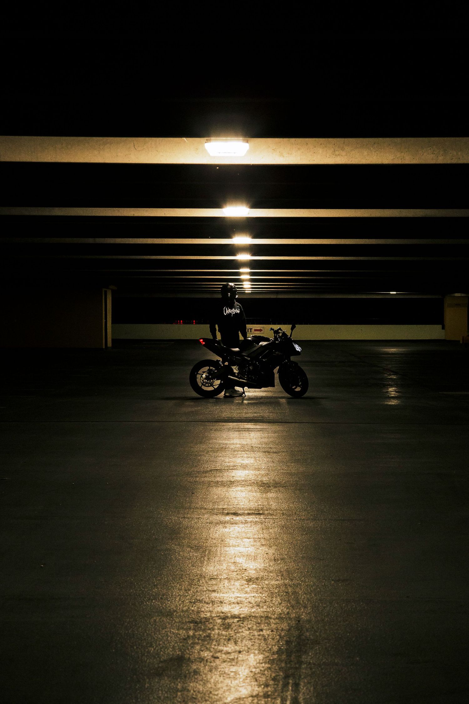 Lone motorcyclist in dark gear sits on a sport bike in a dimly lit parking garage, with overhead lights reflecting on the concrete floor.