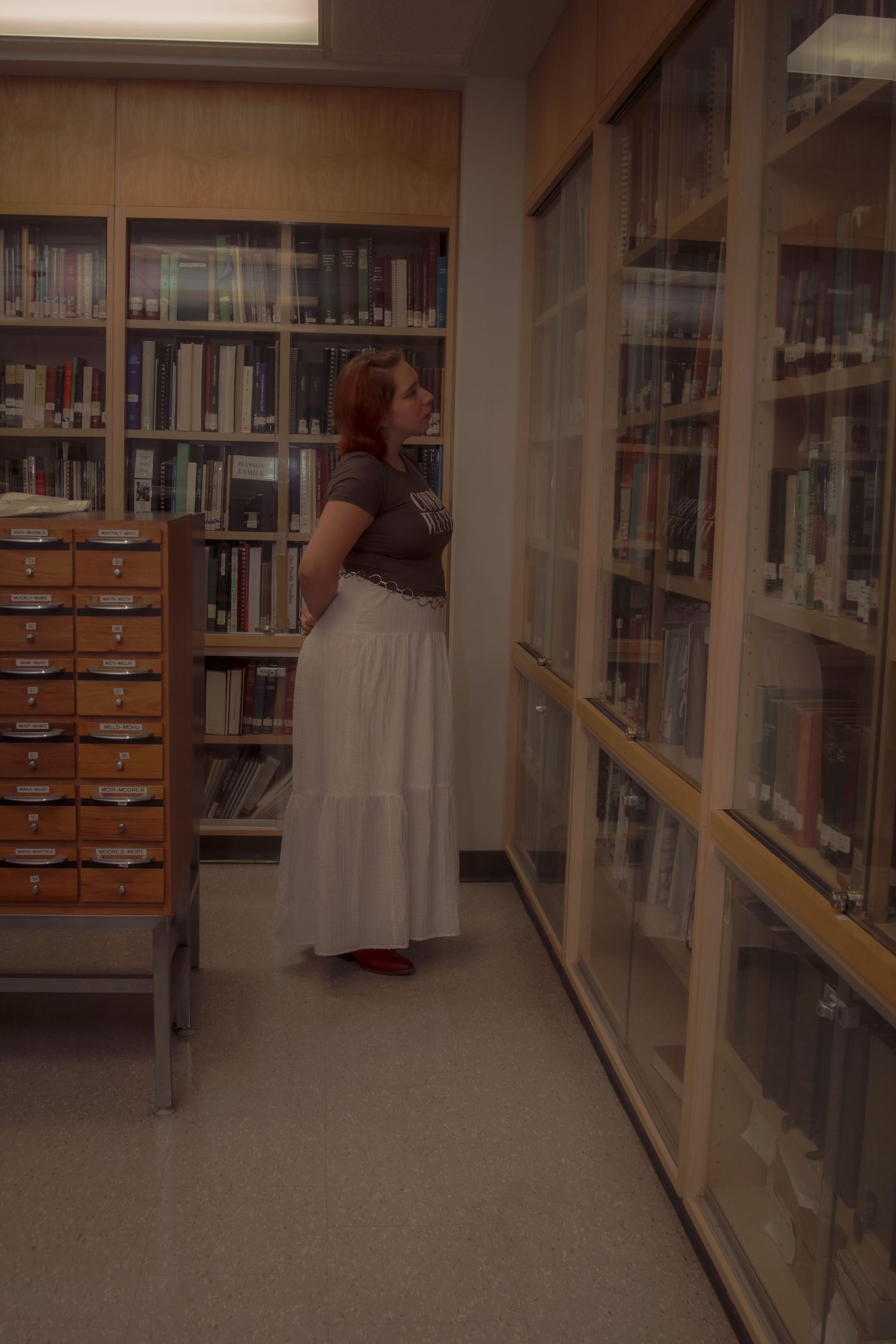 A person browses books in a library aisle with wooden shelves and fluorescent lighting.