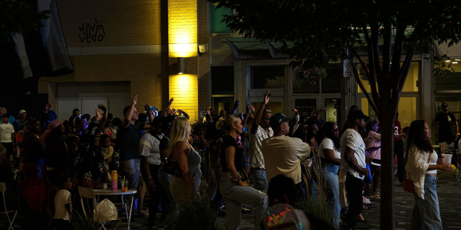 Crowd of people gathered outdoors at night near illuminated building with trees in urban setting.