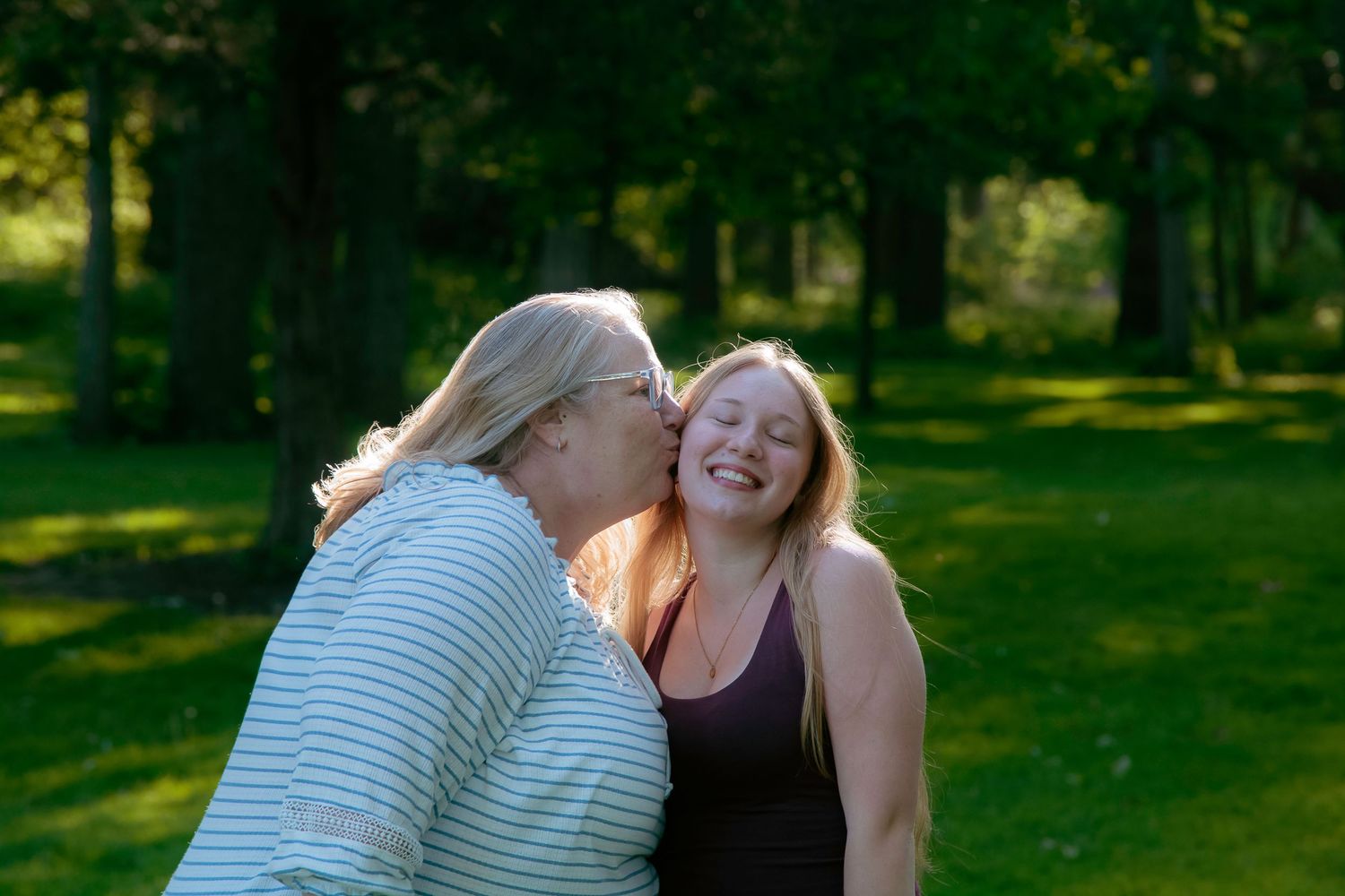 An older woman kisses the cheek of a smiling young woman outdoors in a lush green park setting.