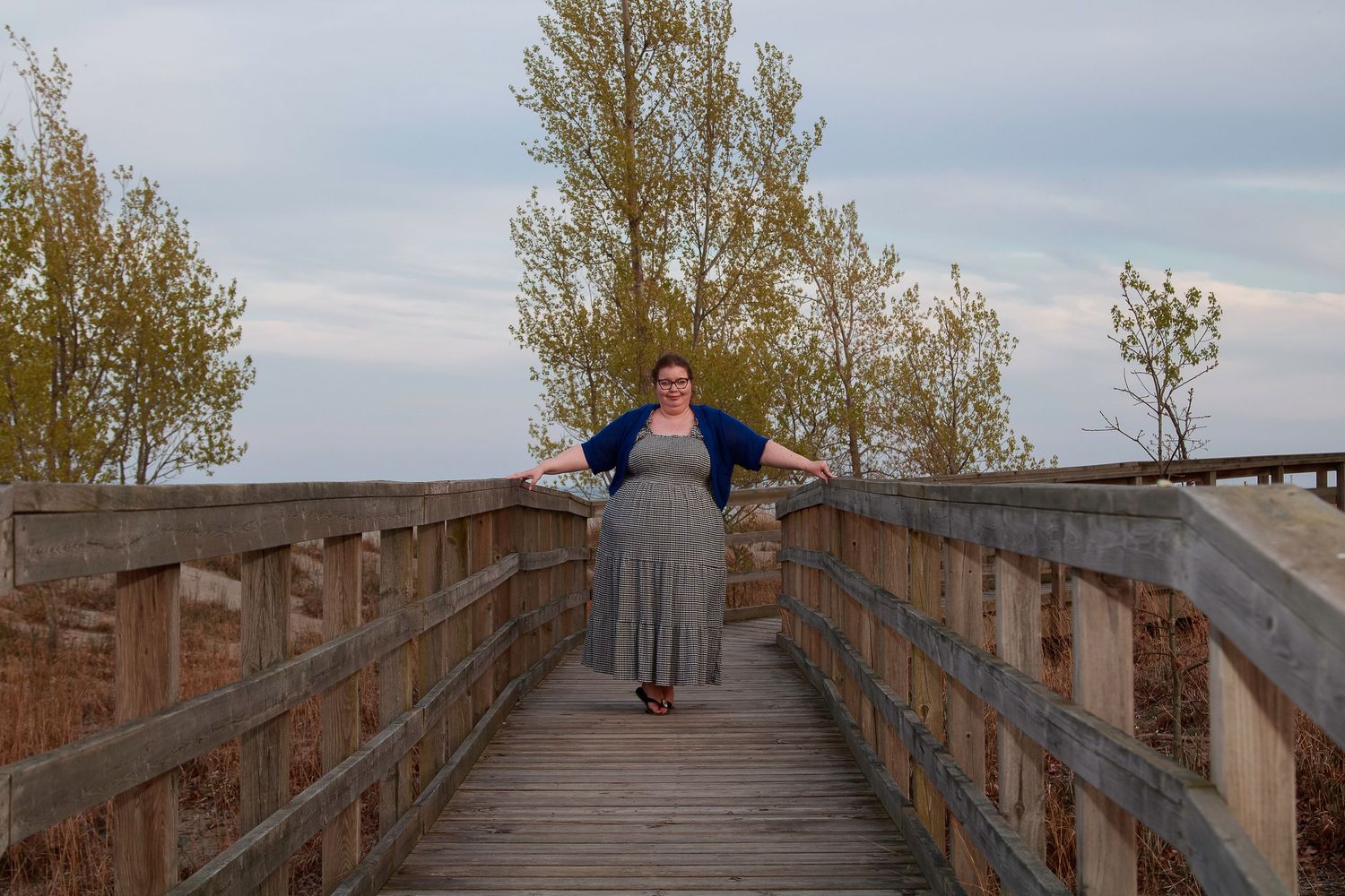 Woman in gray dress and blue cardigan poses on wooden boardwalk surrounded by trees under cloudy sky.