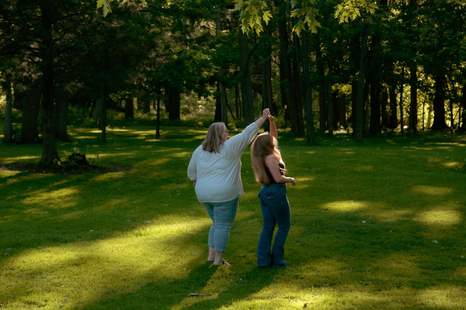 Two women dancing together on lush green grass surrounded by tall trees in a sunny outdoor park setting.