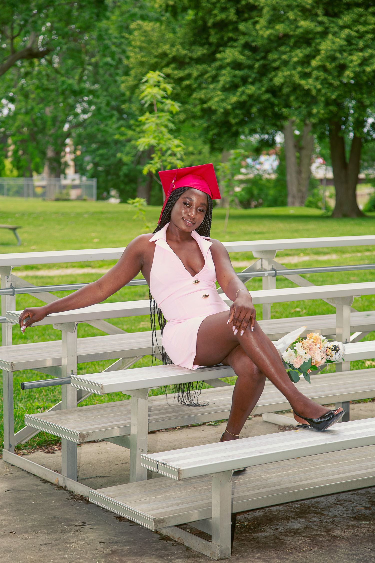 Young Black woman in pink dress and red graduation cap poses confidently on bleachers outdoors holding a bouquet.