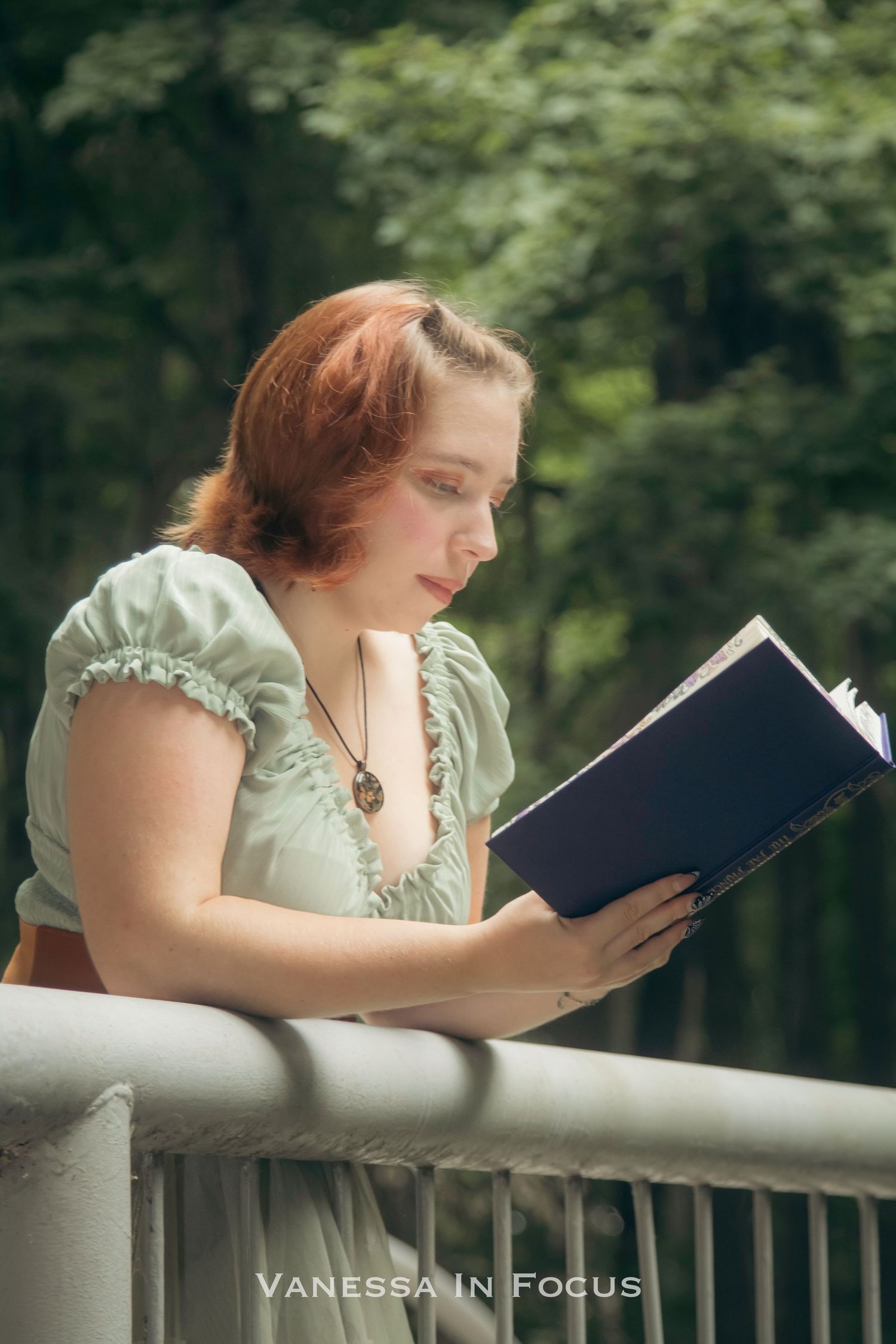 A person in a mint green dress reads a book while leaning on a white railing in a garden setting.