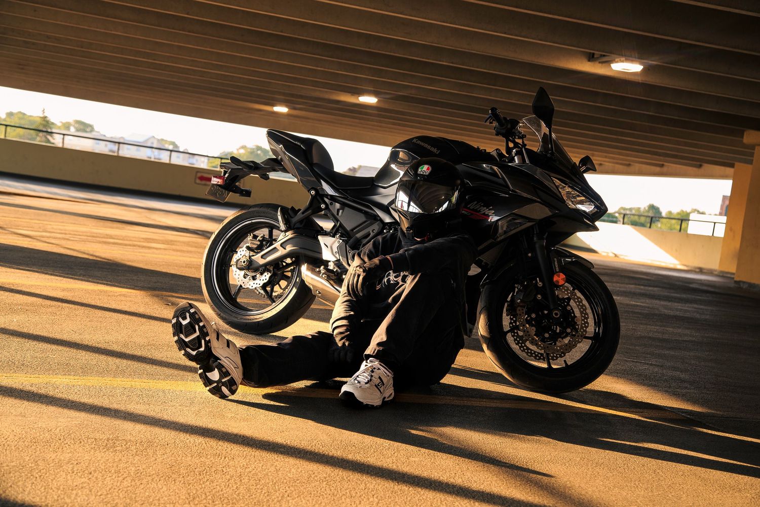 Black sport motorcycle parked under a bridge overpass with a rider crouching beside it in full riding gear.