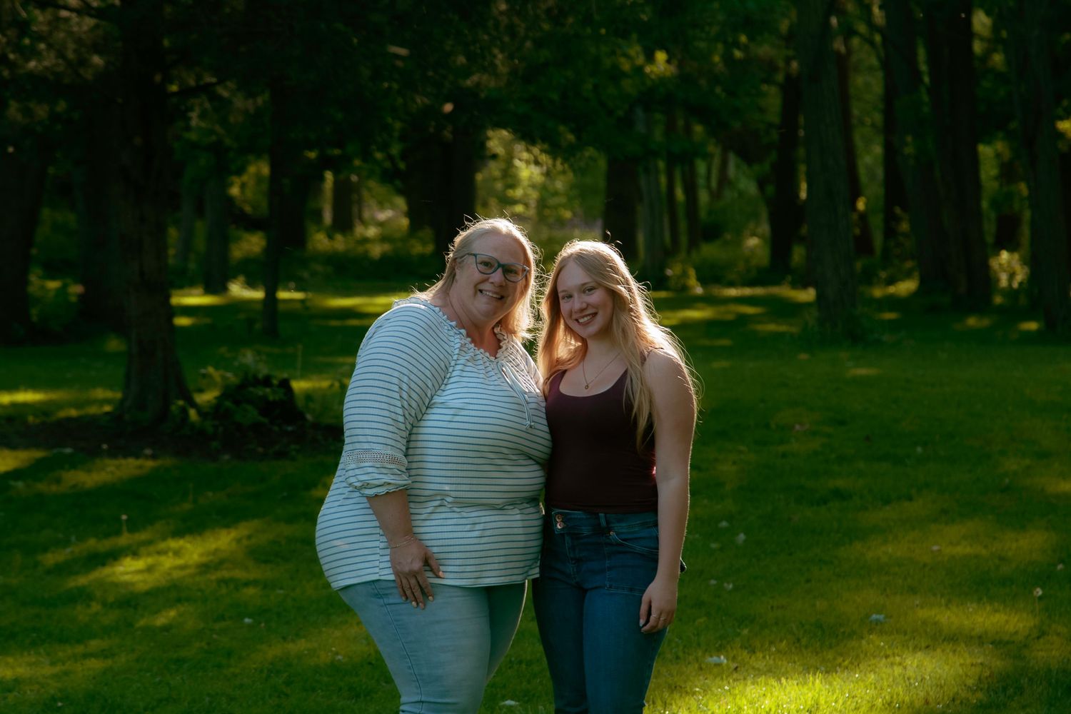 Two women smiling together outdoors in a sunlit green park surrounded by tall trees.