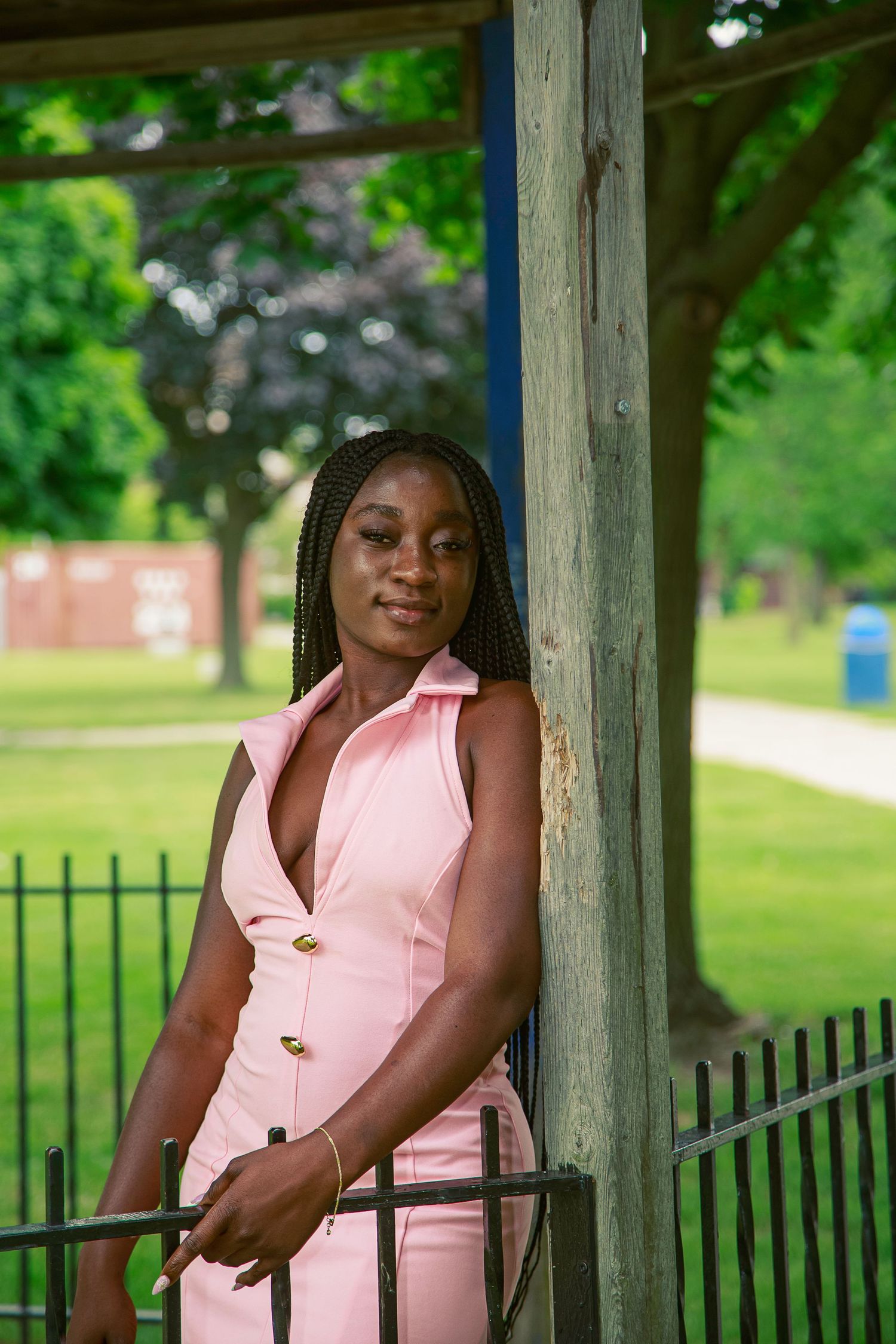 A young Black woman in a pink sleeveless jumpsuit poses gracefully by a wooden pergola post in a lush green park.