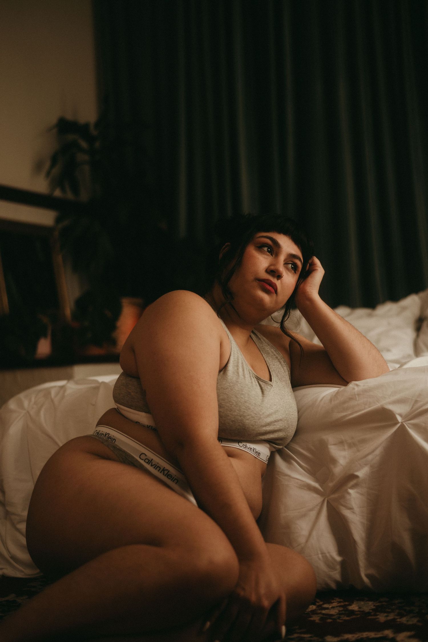 Moody boudoir photograph of a person posing on white bedding in dim lighting.