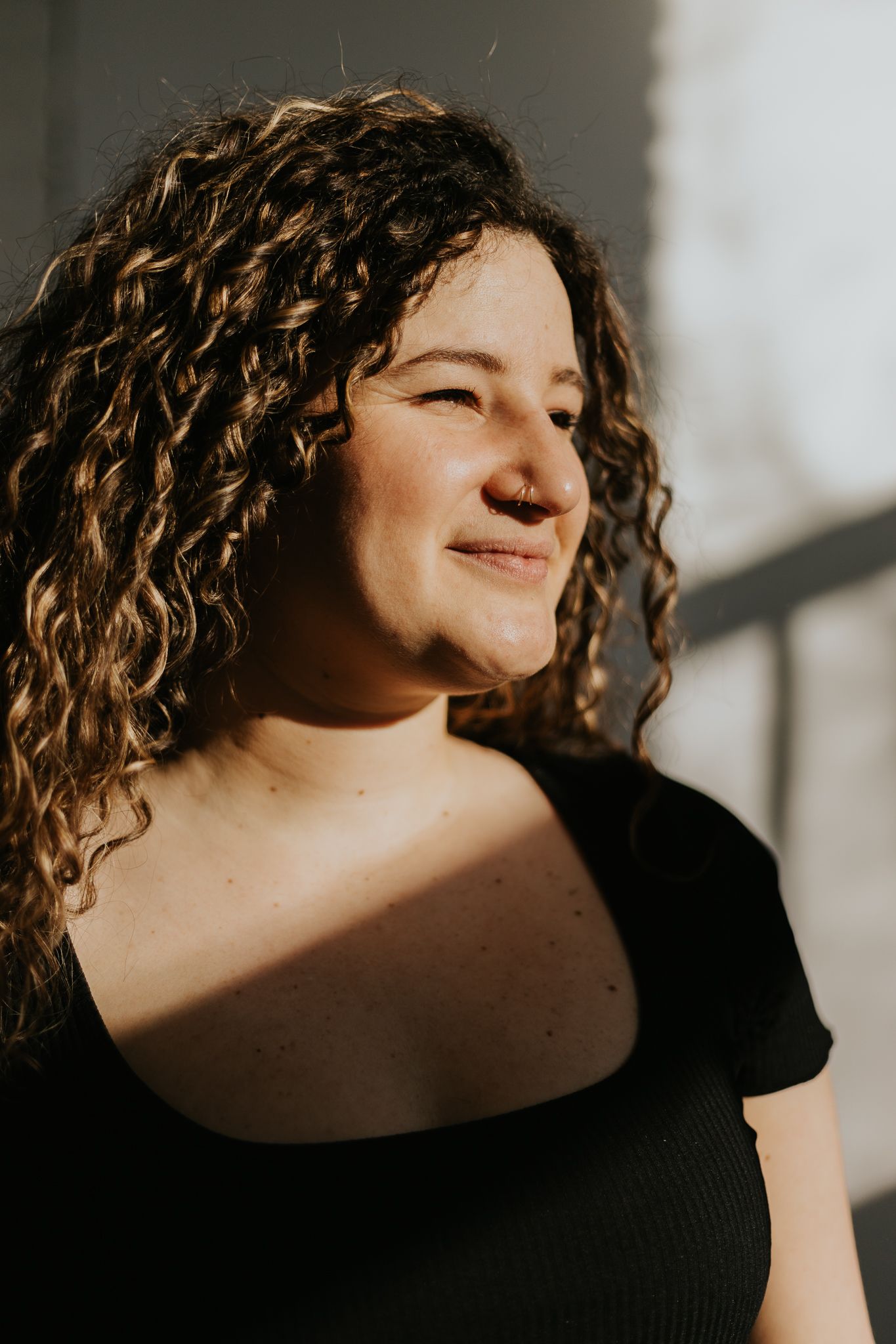 Portrait of someone with natural curly hair smiling in warm sunlight while wearing a black top.