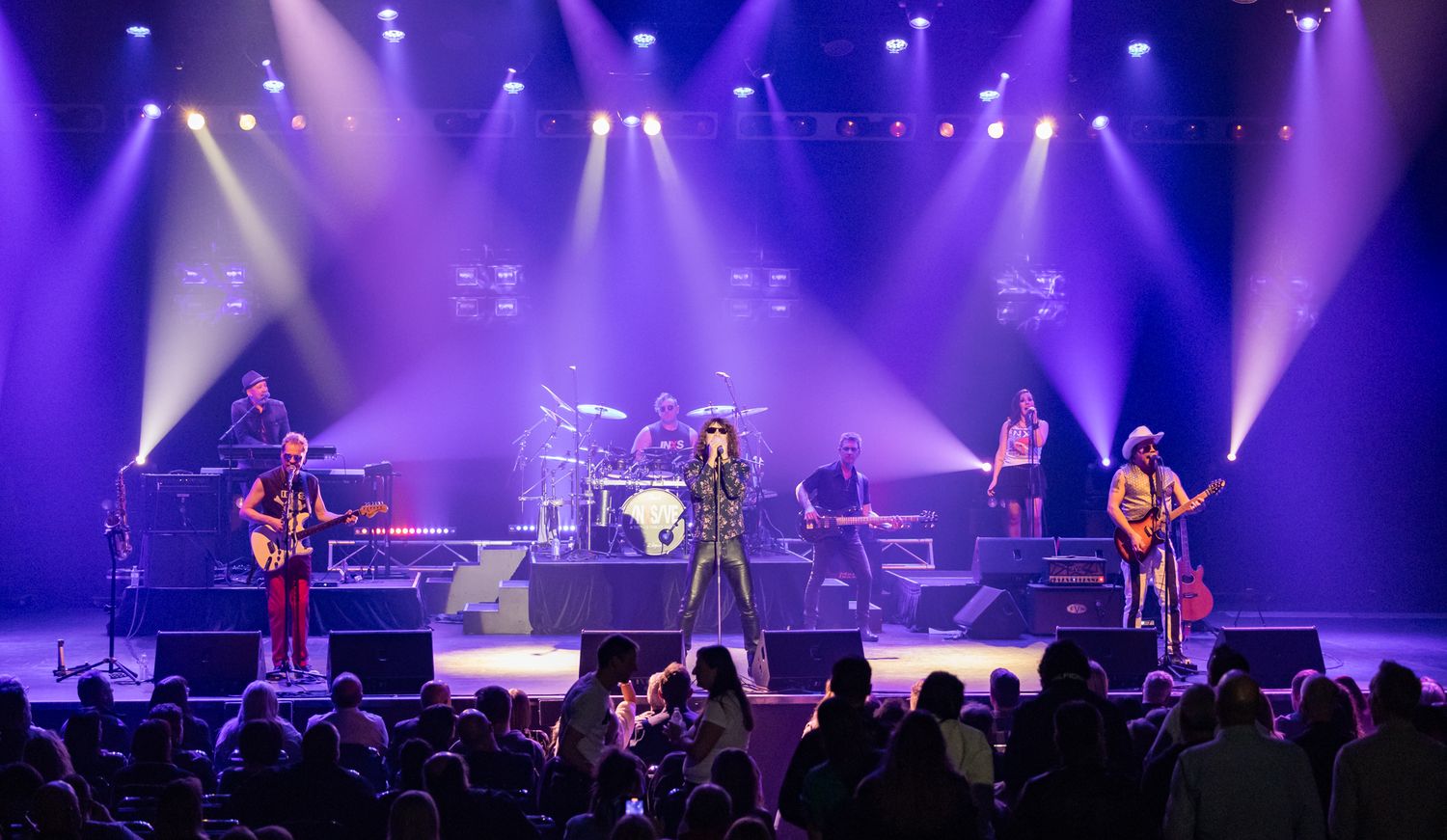 Rock band performing live on a well-lit purple stage with multiple guitarists and a full crowd audience.